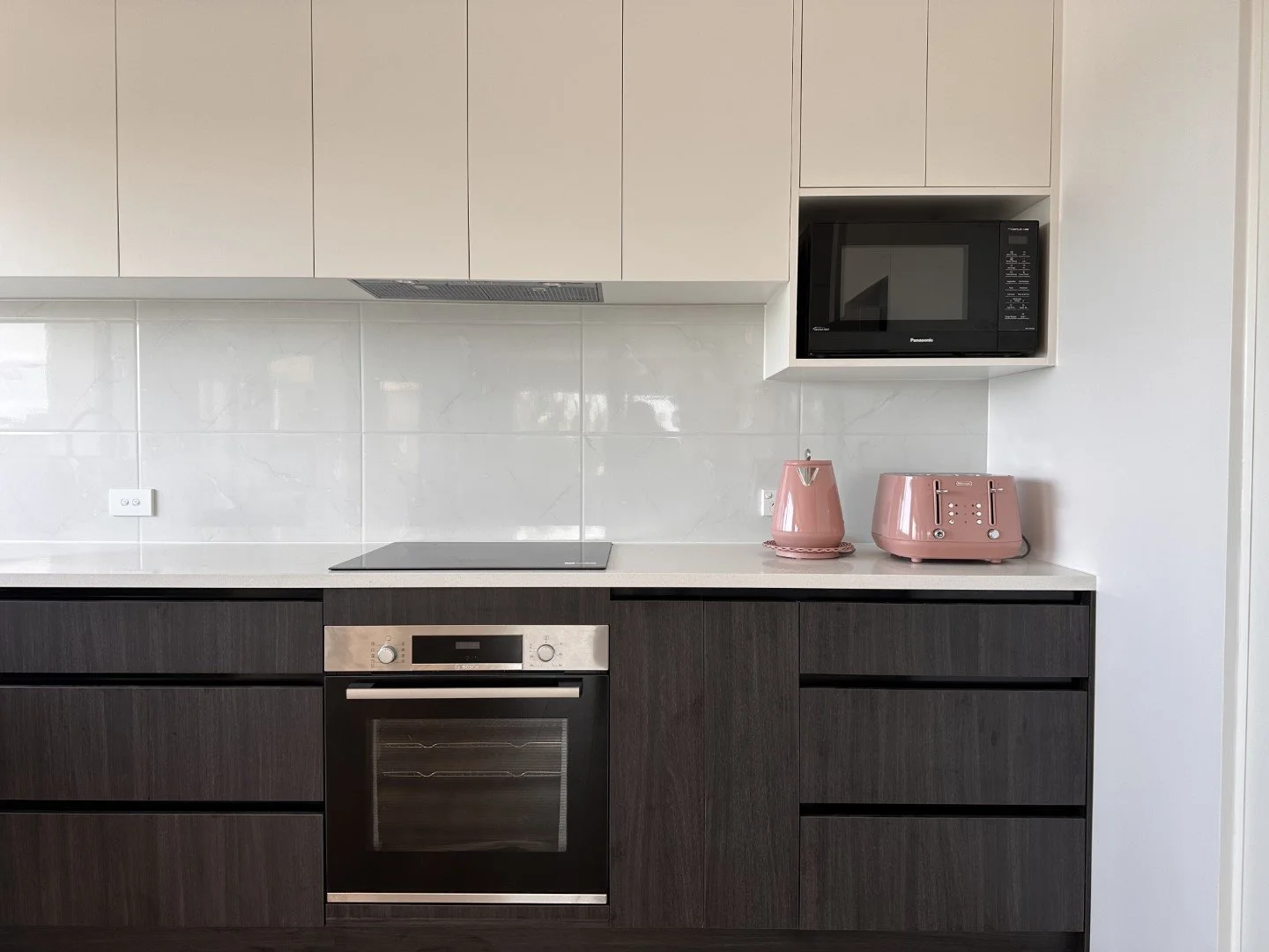 Part of a modern kitchen with white upper cabinets, dark lower cabinets, a built-in oven, a black microwave in a wall niche, a pink kettle, and a pink toaster on the countertop.