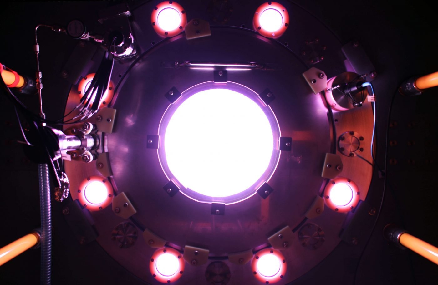 View of a fusion reactor with a bright white circle at the center, surrounded by eight illuminated circular ports in a circular arrangement, featuring various cables, sensors, and mechanical parts.