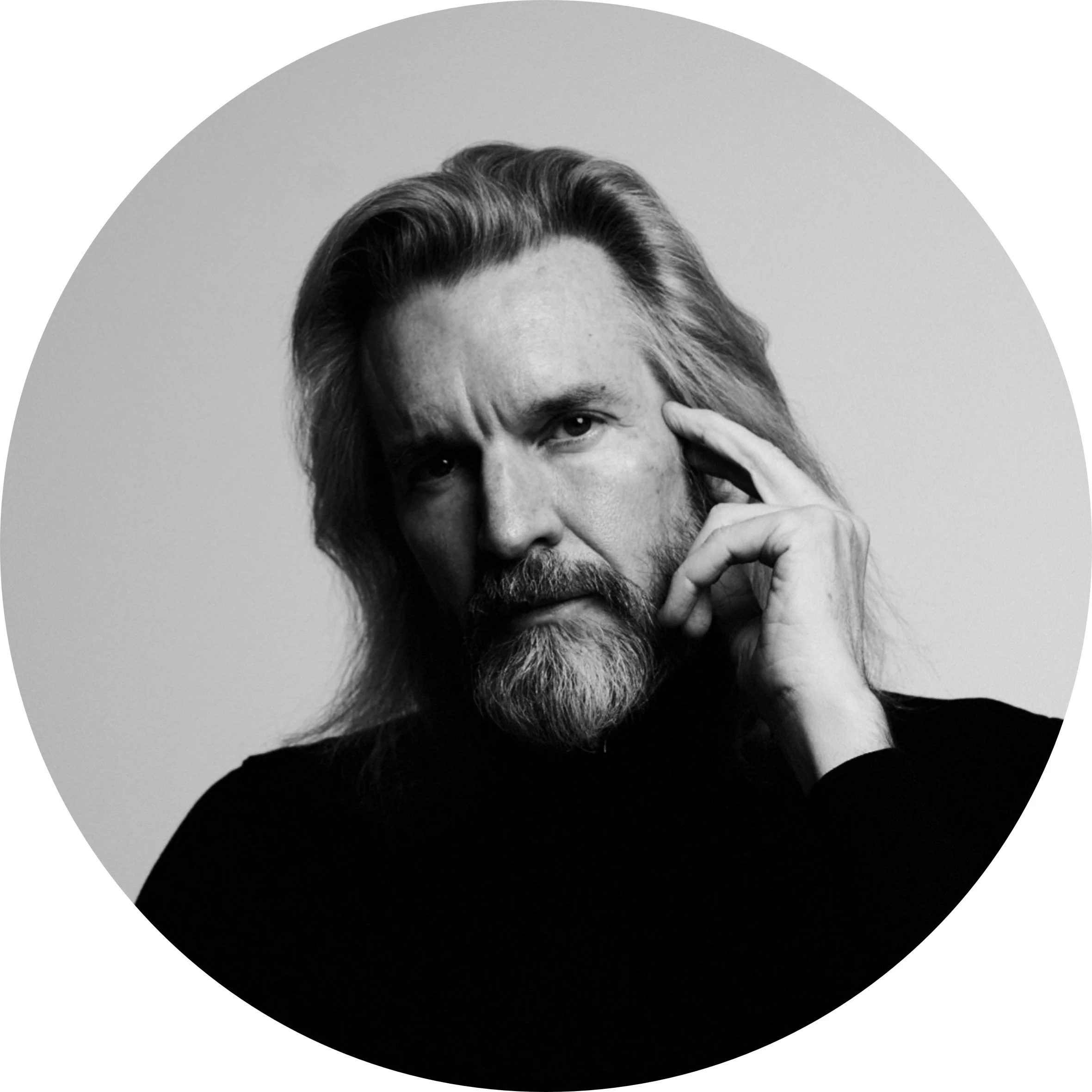 Black and white portrait of a middle-aged man with long hair and a beard, touching his temple with his hand, looking serious.