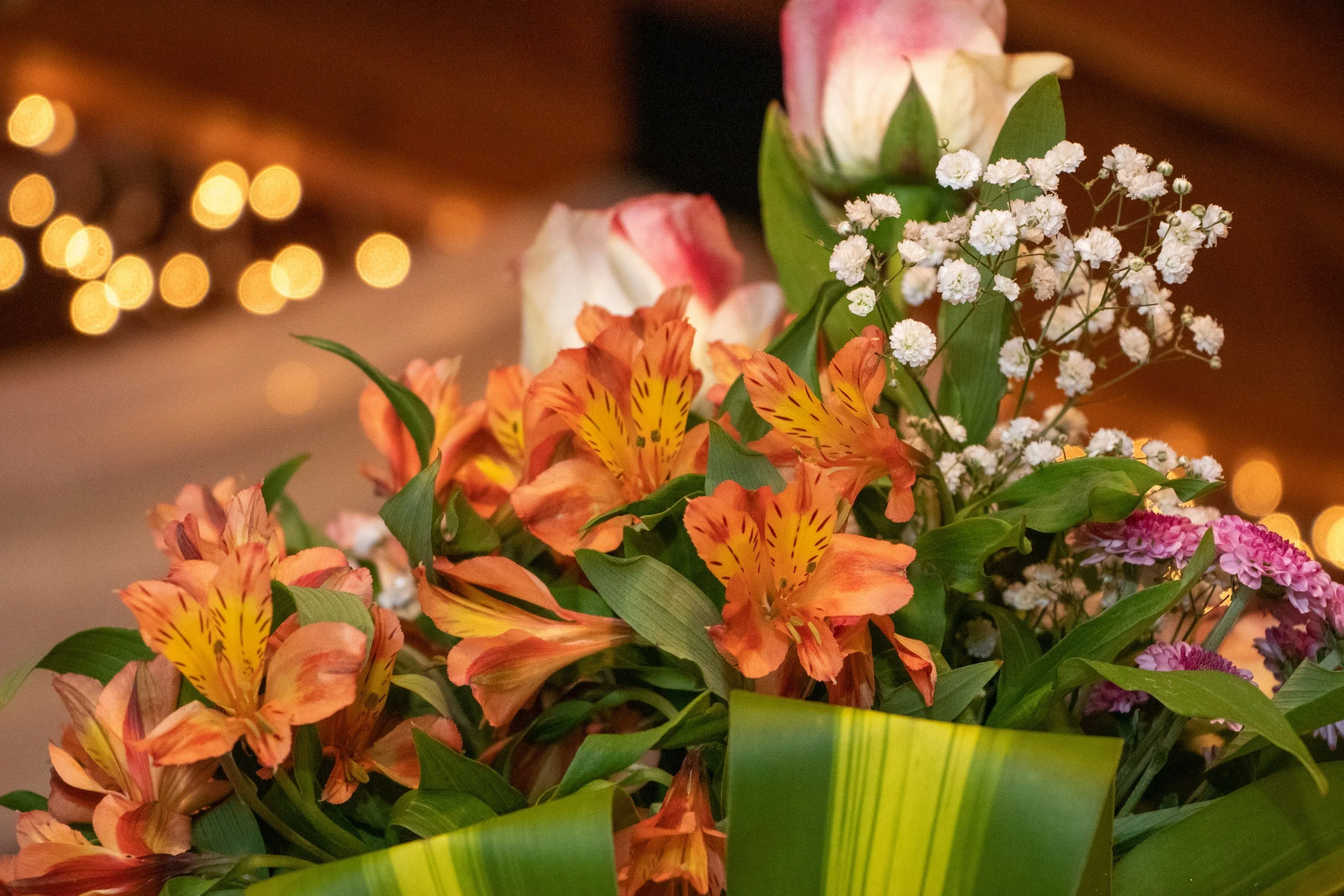 Close-up of a colorful bouquet of flowers including orange and yellow lilies, pink roses, white baby's breath, and purple flowers, with a warm, blurred background and string lights.
