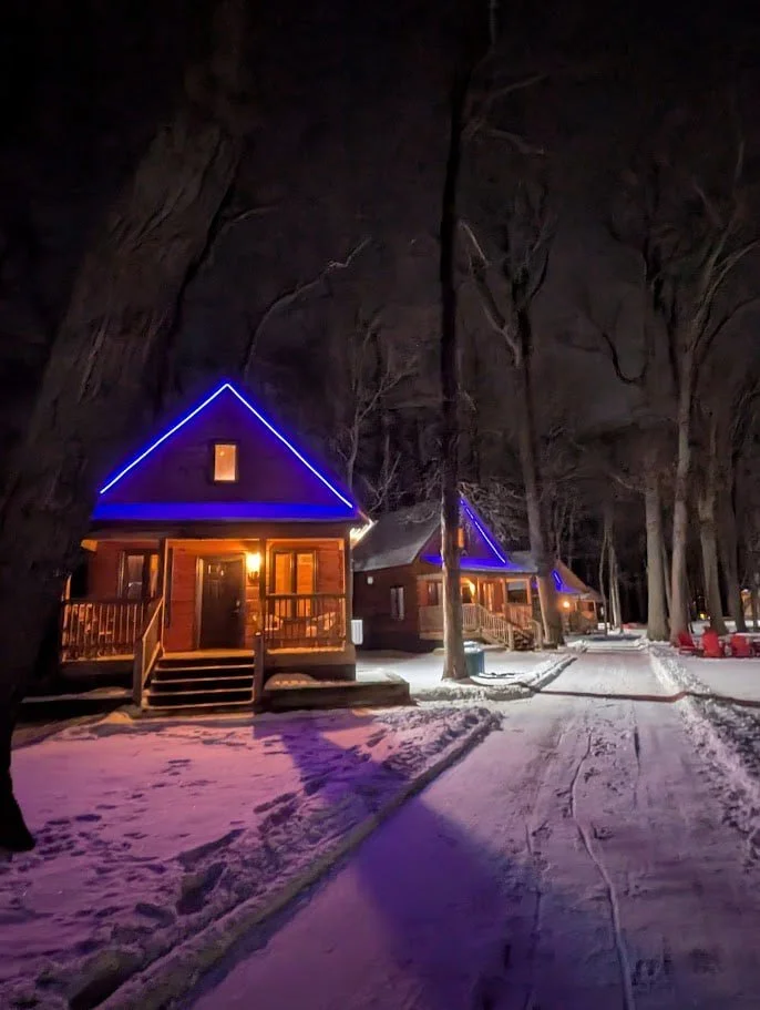 pictures of cabins in the dark, with bright lights in blue on the outstide of the cabin.  it looks like winter as there is white snow on the ground.