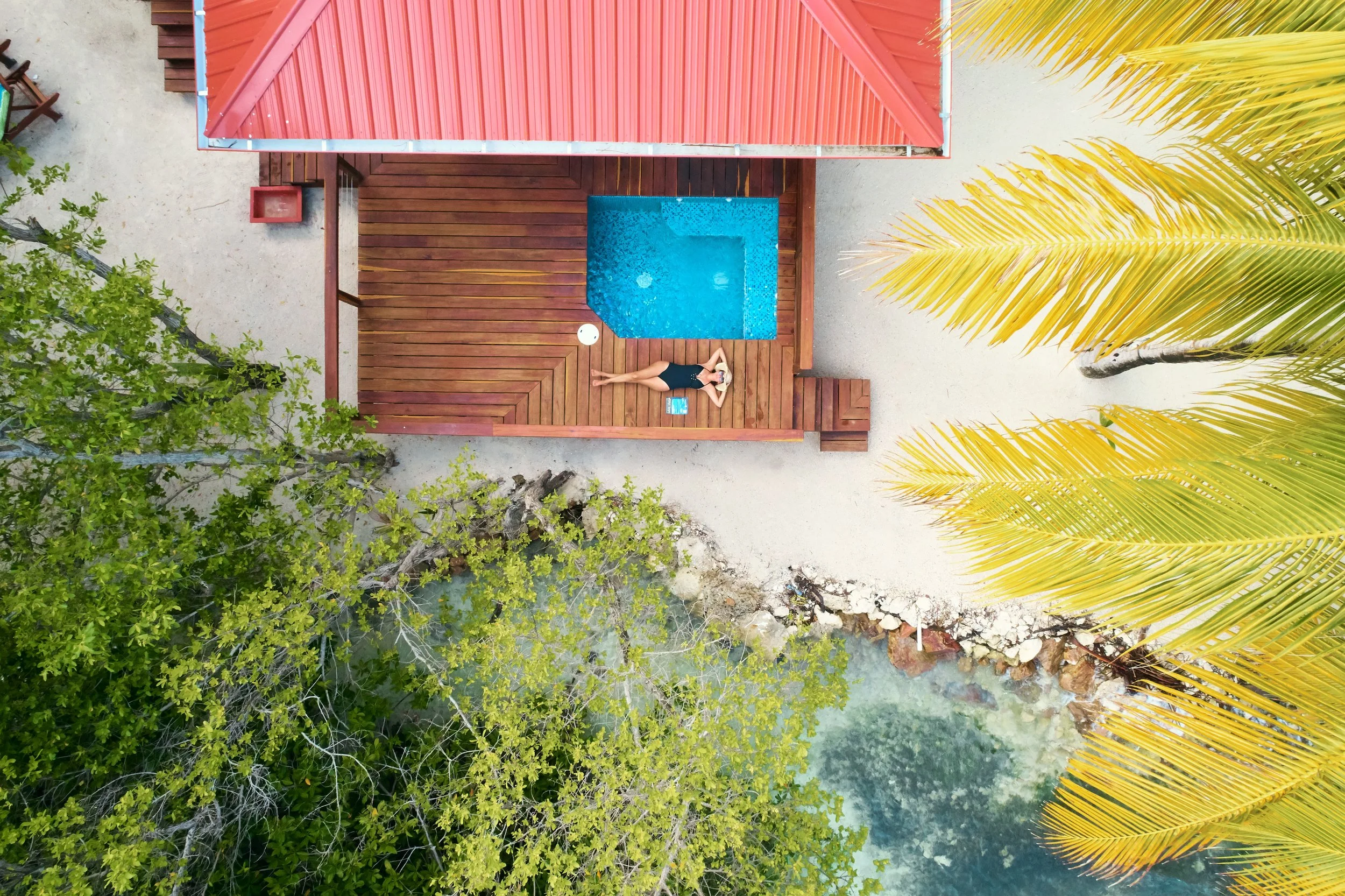 A woman in a black swimsuit lying on a wooden pool deck near a small swimming pool, surrounded by green trees and palm fronds.
