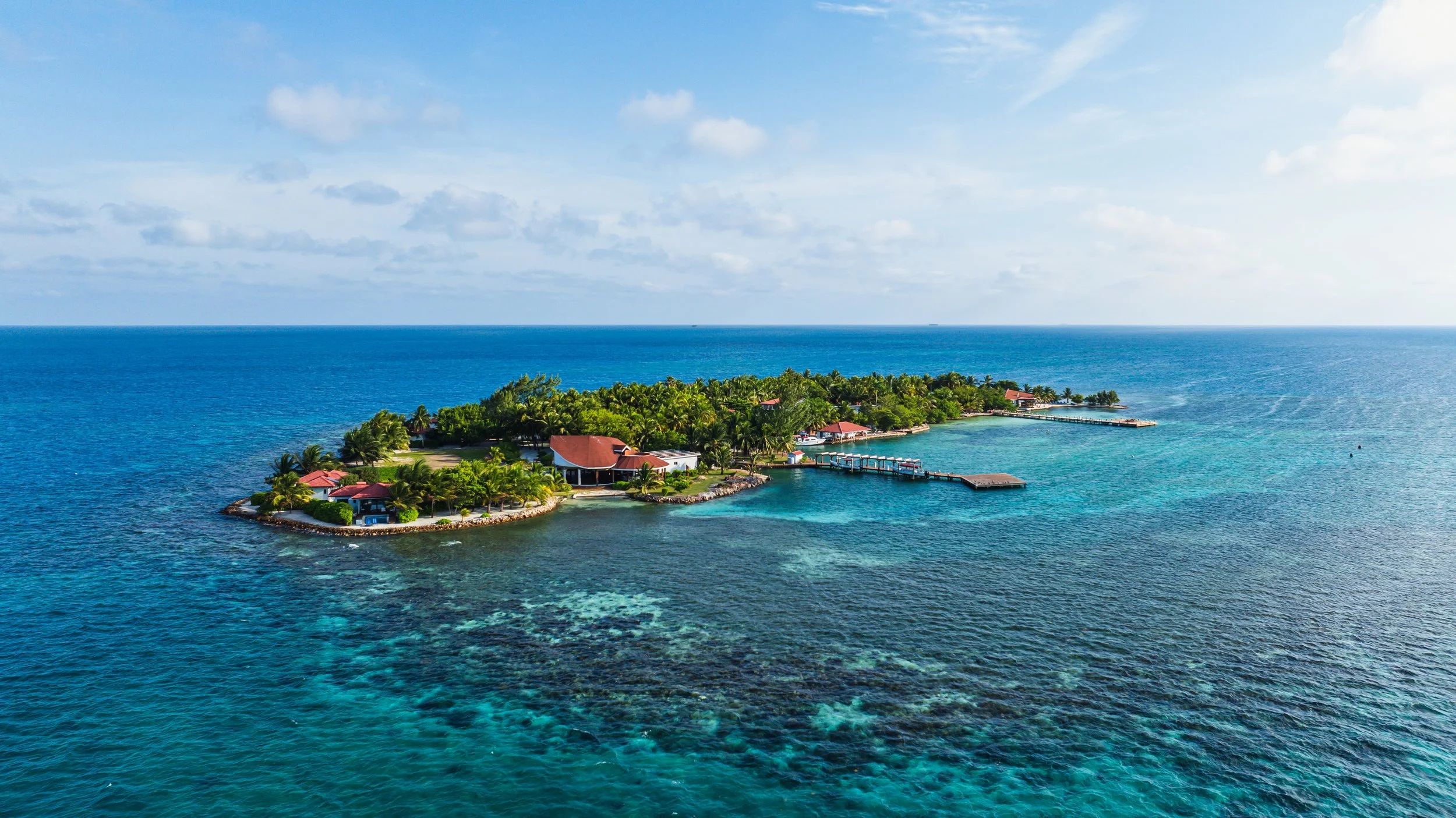 Aerial view of a tropical island with lush greenery, several buildings with red roofs, and docks extending into the turquoise ocean. The sky is partly cloudy.