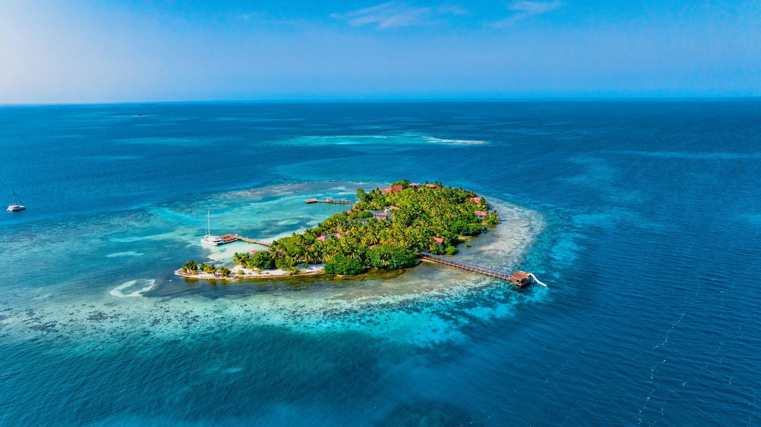 Aerial view of a tropical island with lush greenery, several buildings, a long pier extending into the ocean, sailboats, and turquoise waters surrounding the island.
