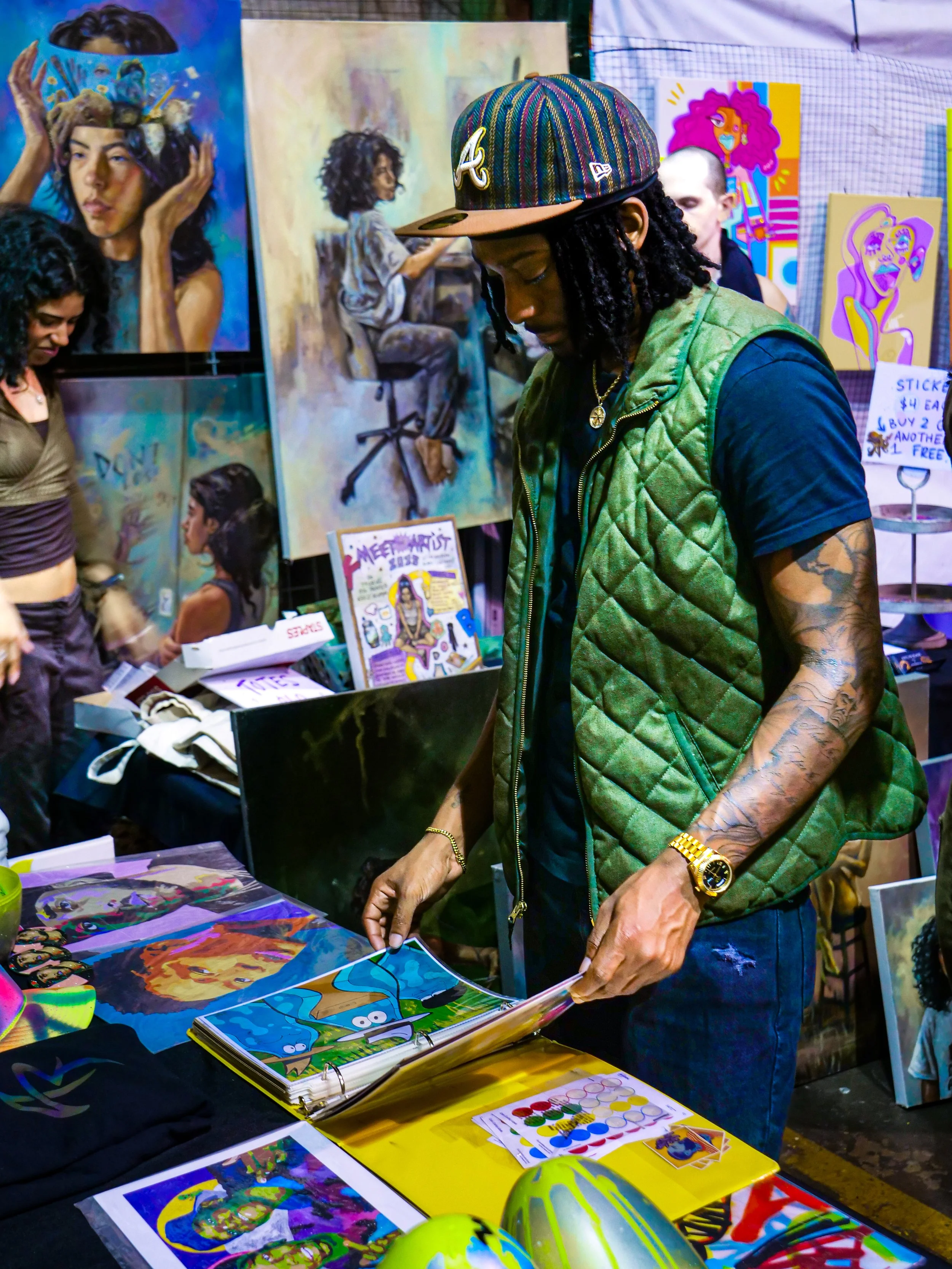 Three women are engaged in conversation at an art exhibition featuring colorful paintings and illustrations, including an anime-style artwork. The woman on the left has long, wavy hair with blonde highlights, is wearing a denim jacket and a white, tiered skirt, and carries a beige tote bag. The woman on the right has short black hair, wears a black crop top, camouflage pants, a striped knit hat, and has a drink in her hand. The background shows a brick wall with various artworks displayed.