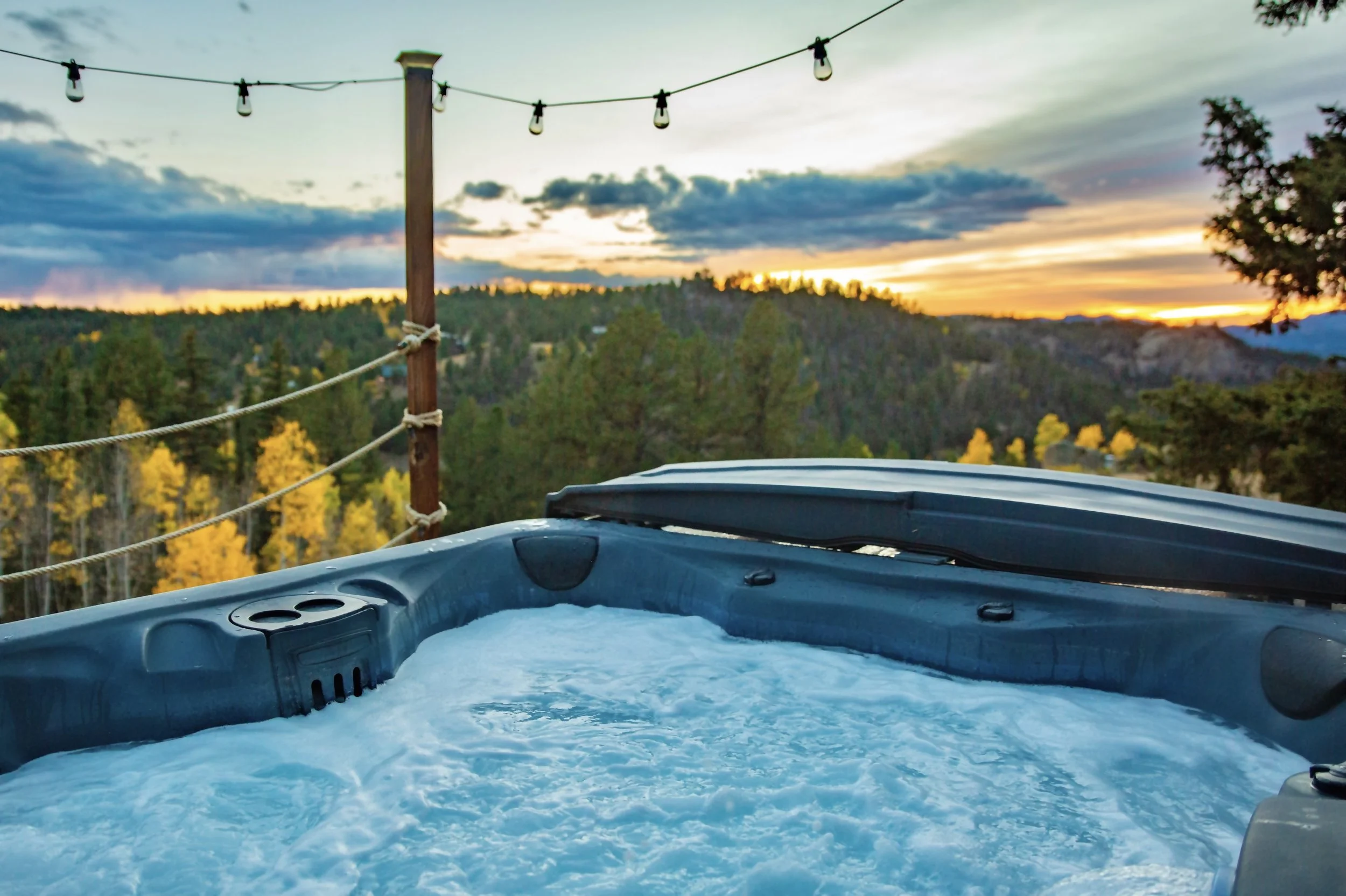 A hot tub filled with bubbling water outdoors at sunset, overlooking a forest with trees showing fall colors, and a cable line with lights strung across the scene.