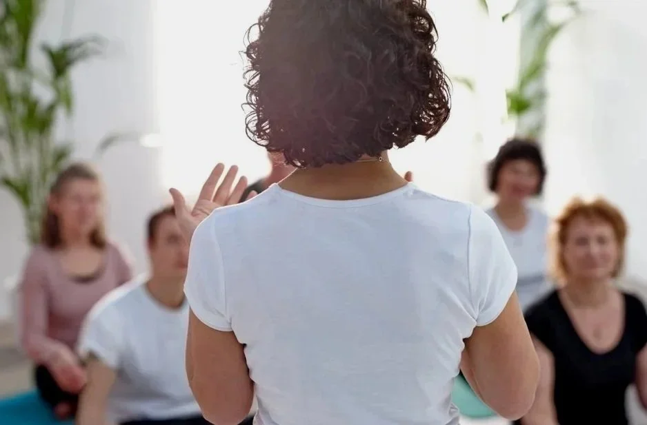 A woman with curly hair leading a group in a discussion or presentation in a bright, indoor setting with plants in the background.