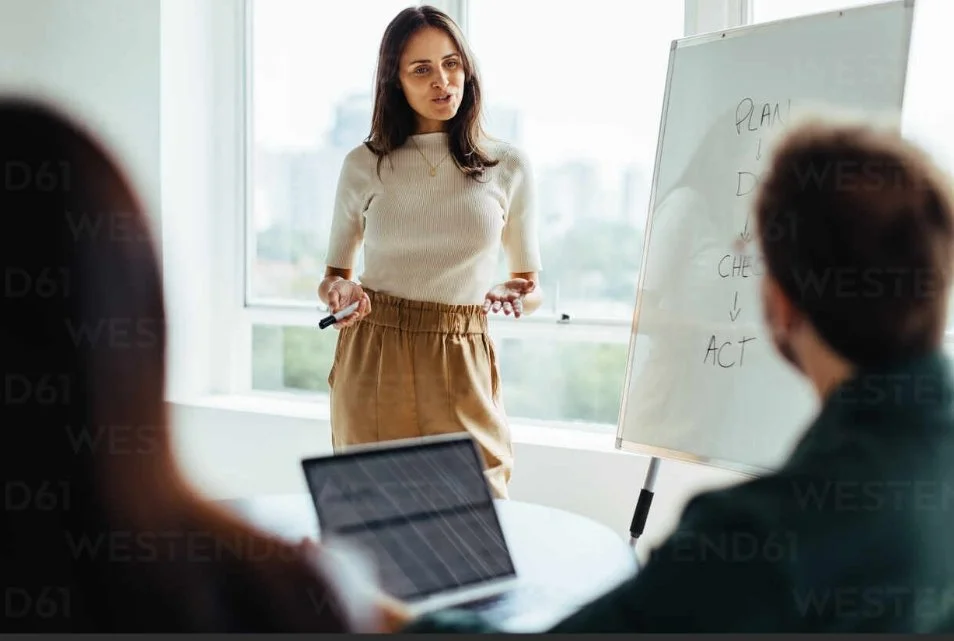 Woman giving a presentation to a small group in a bright office, standing near a whiteboard with notes, while others work on laptops.