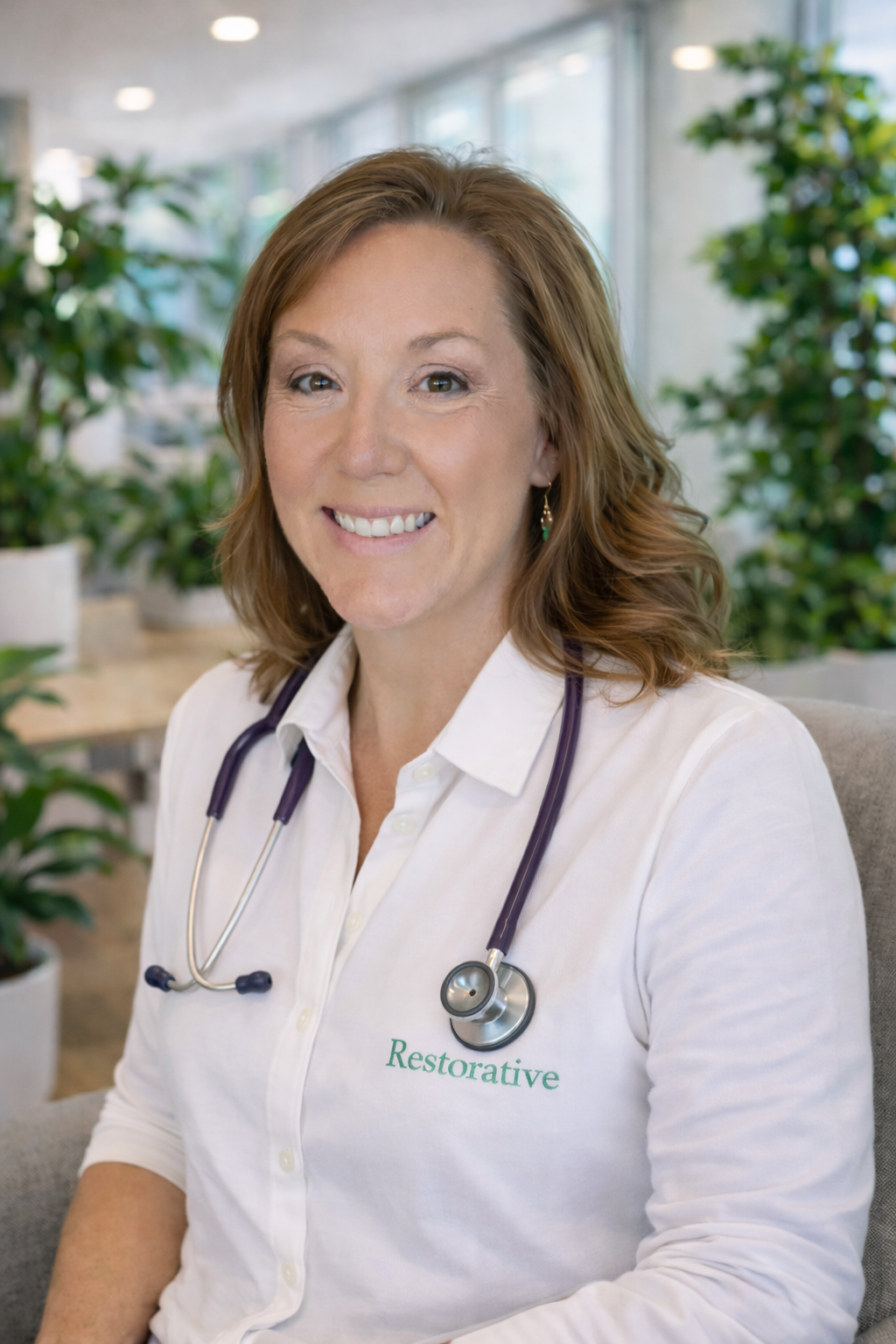 A smiling female healthcare professional with a stethoscope around her neck, wearing a white shirt with 'Restorative' embroidered on it, sitting in a bright office or medical setting with plants in the background.