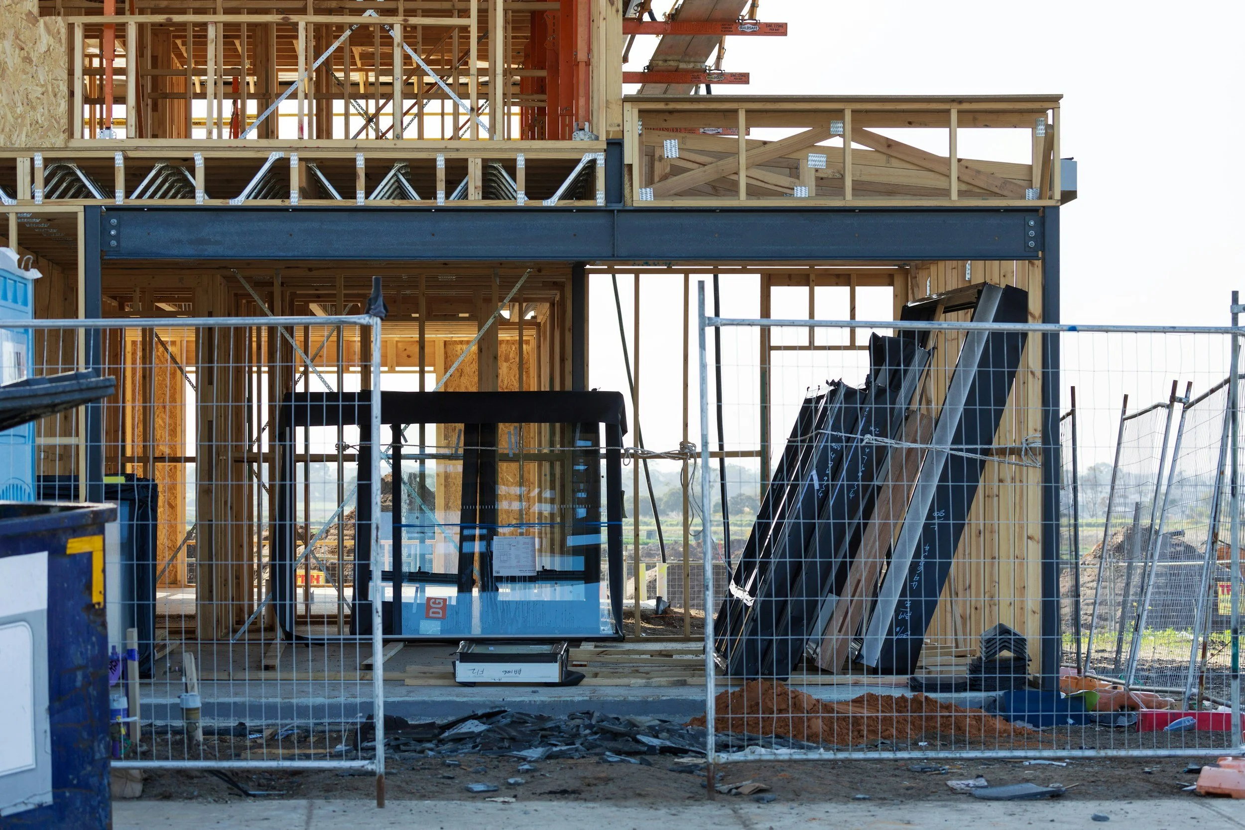Construction site of a multi-story building with exposed wooden framing and scaffolding, surrounded by temporary fencing.