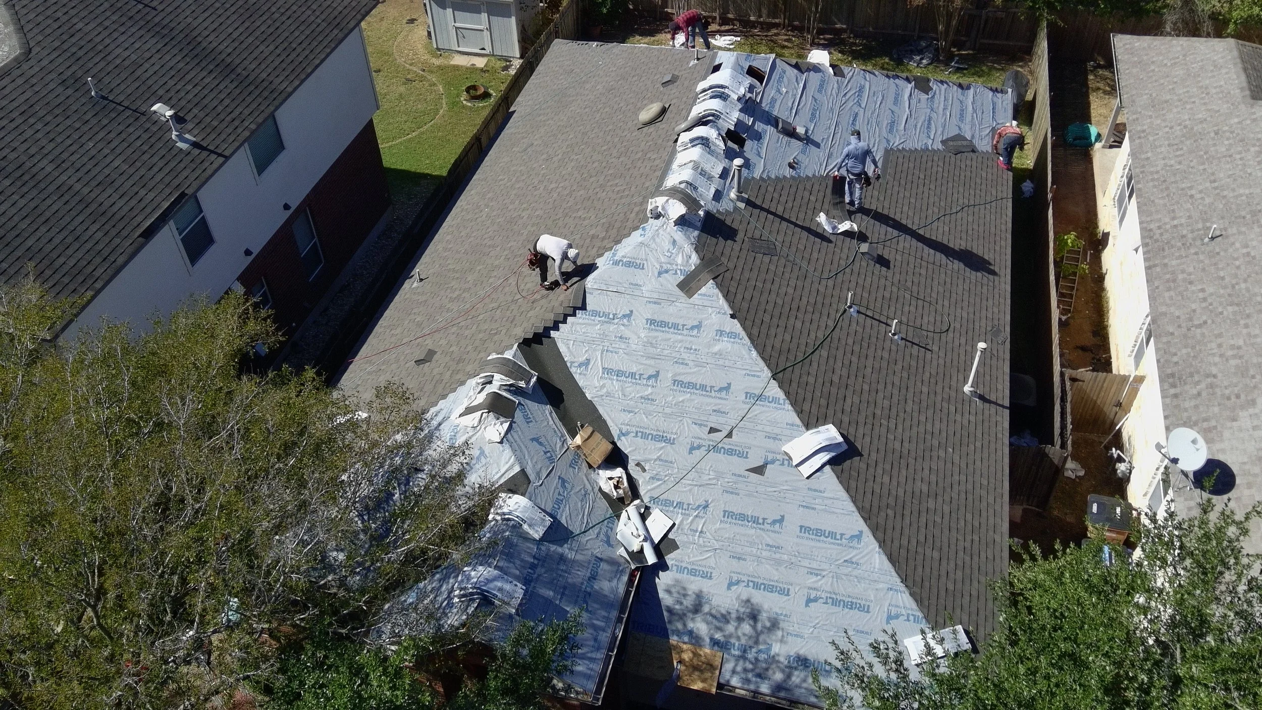 Aerial view of workers repairing the roof of a house in Lakeway Austin, with some sections covered in a silver underlayment and others with roofing shingles, surrounded by neighboring houses and trees.