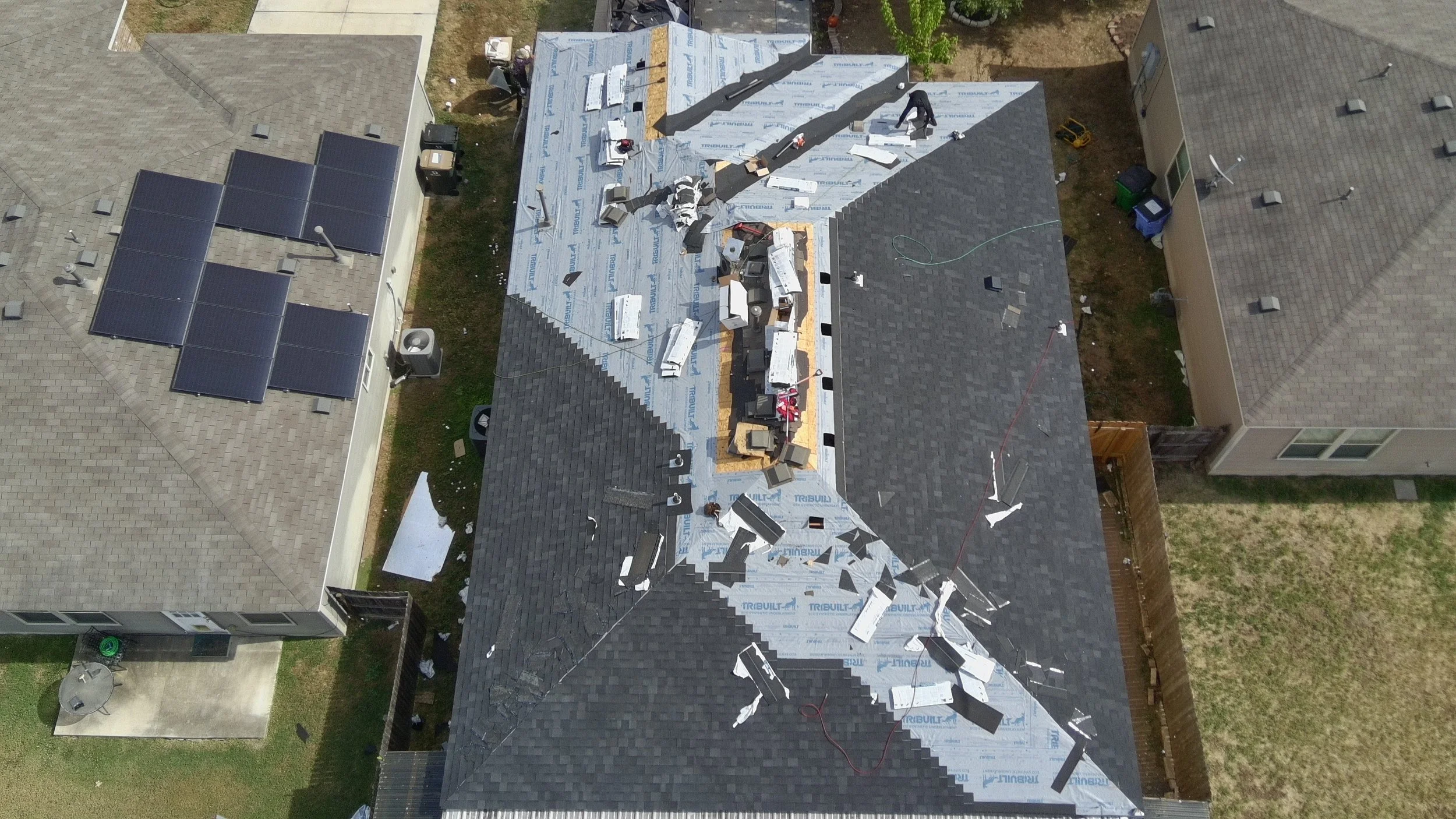 Aerial view of a house in Leander, roof under construction with roofing materials, tools, and workers on top. Solar panels are installed on the neighboring house's roof.