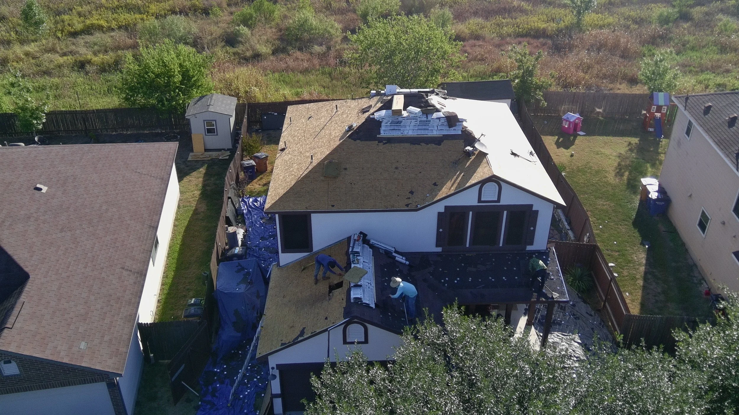 Aerial view of a house in Round Rock, Austin, under renovation with workers on the roof and yard, neighboring houses, a garden with children’s play equipment, and trees in the background.
