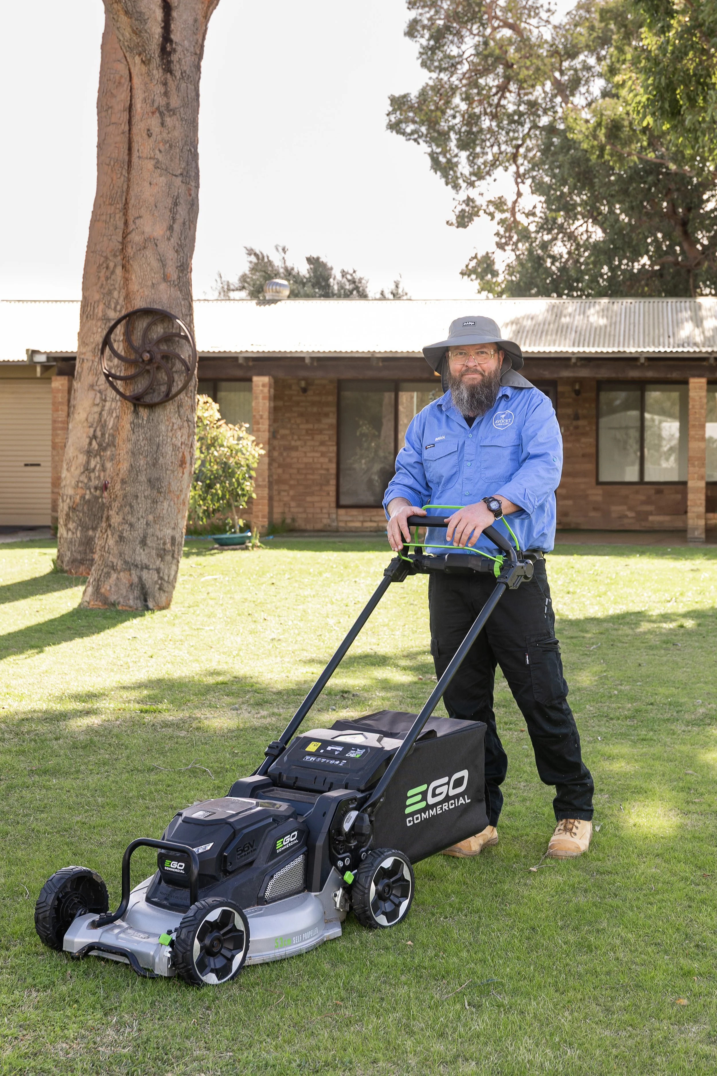 A man with a beard, stands outside looking happy on a grassy lawn, holding a battery lawn mower, ready to offer the best lawn mowing service to people in Wannanup and surrounding areas.