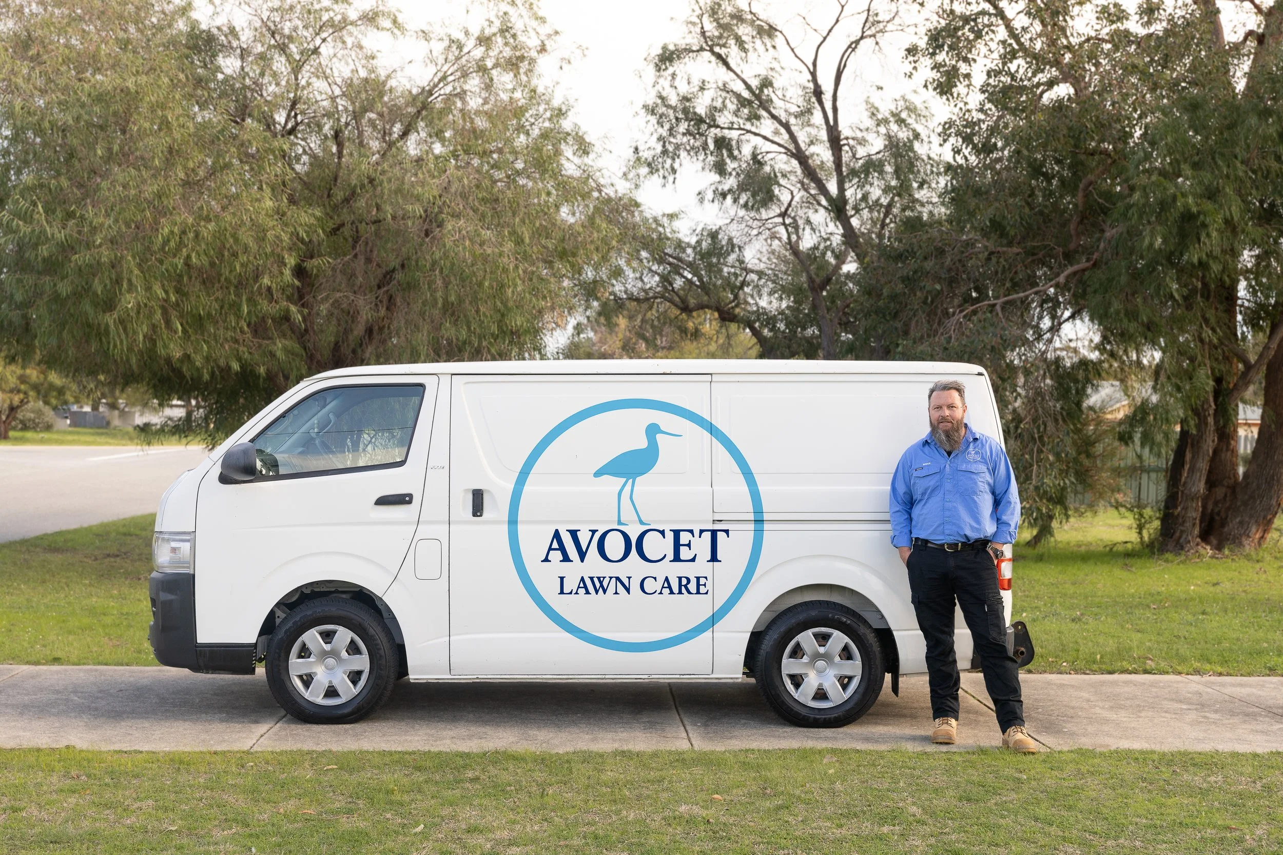 A man standing next to a white van with a logo that reads "AVOCET LAWN CARE" and features a bird illustration, ready to maintain premium lawns and gardens.
