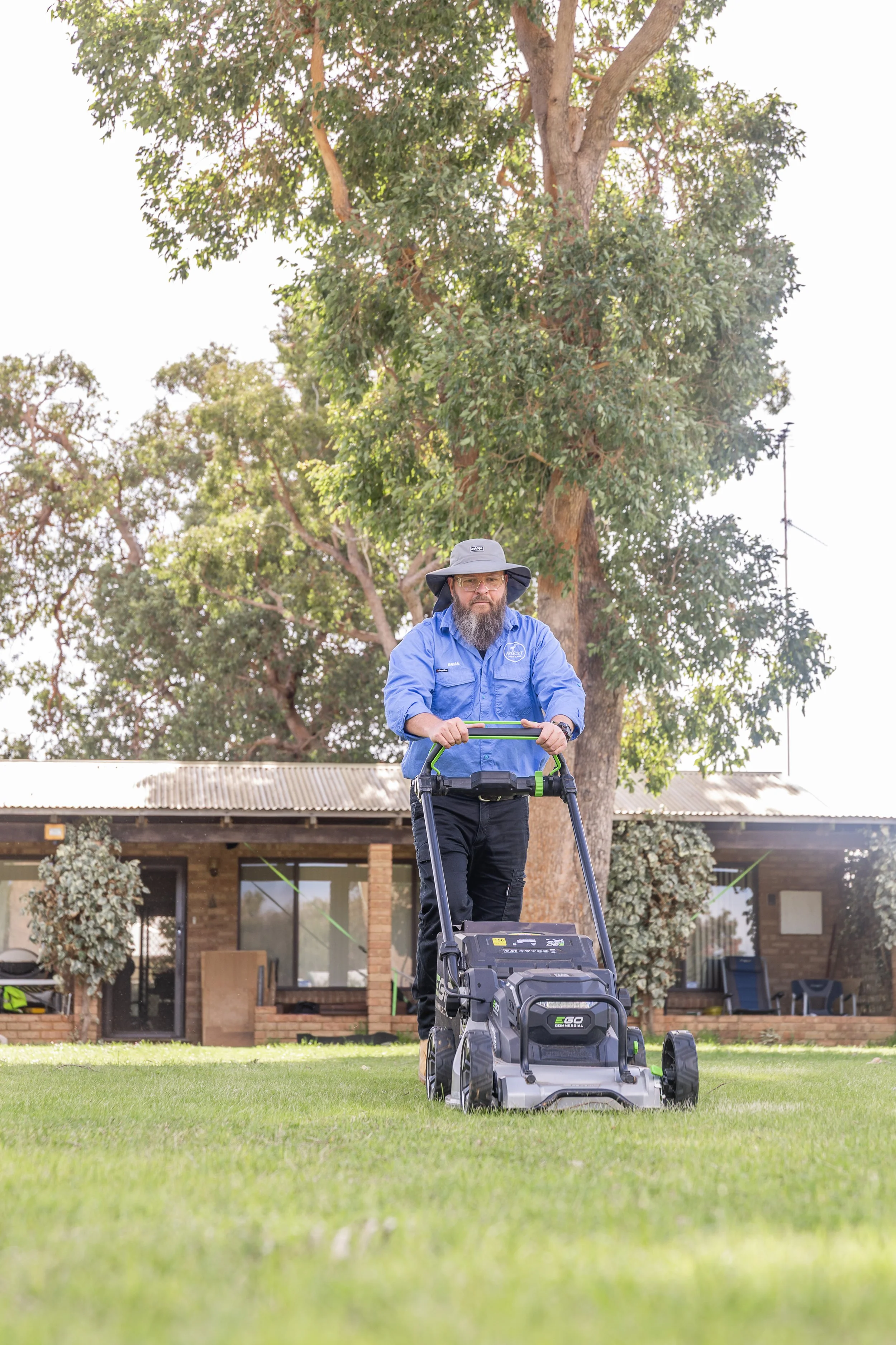 A man with a beard wearing a blue shirt, black pants, and a wide-brimmed hat mows the lawn with an electric lawnmower in front of a house with trees and shrubs.