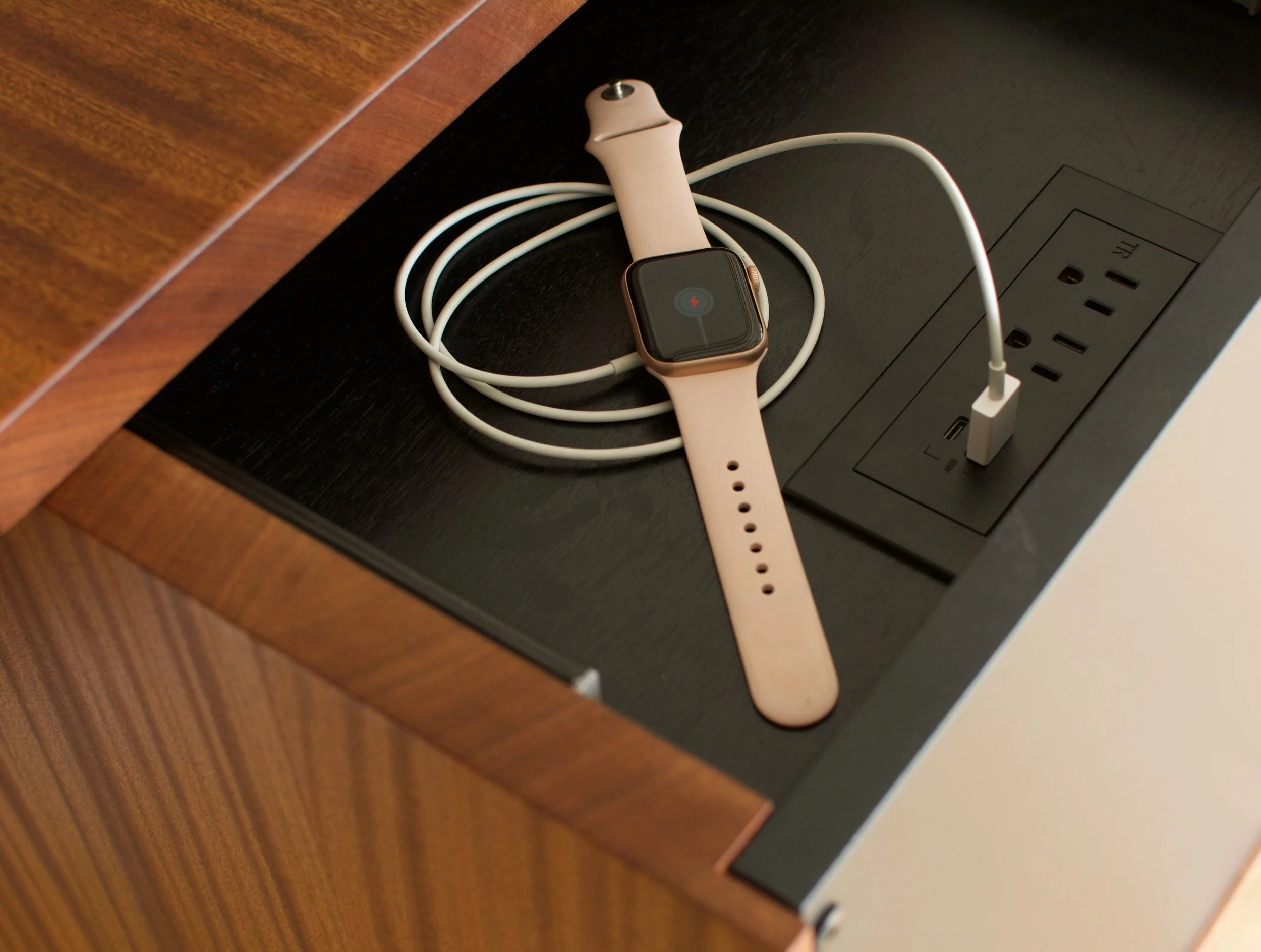 An Apple Watch with a pink band connected with a charging cable on a black wooden desk near a power outlet.