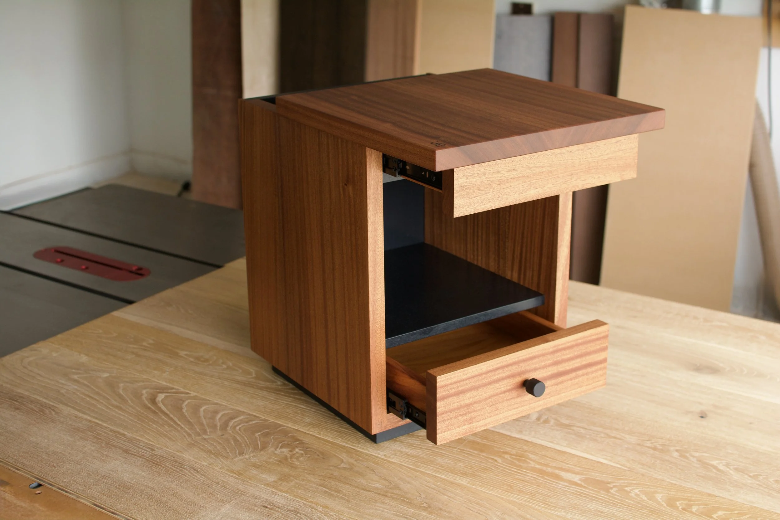 A small wooden cabinet with a drawer and an open shelf, placed on a light wooden table in a workshop or studio.