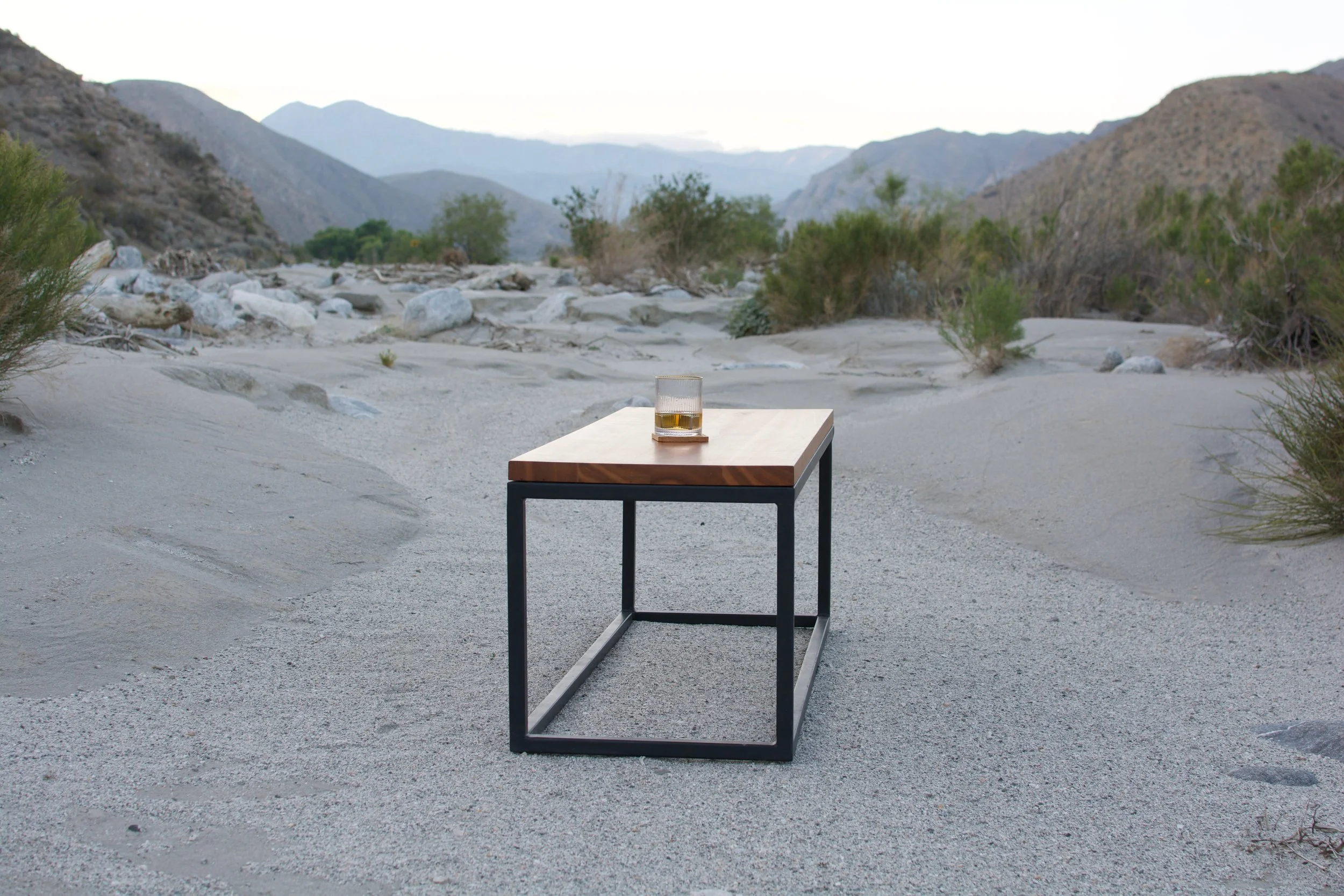 Table with a glass of whiskey in a desert landscape with mountains, sand, and sparse vegetation.