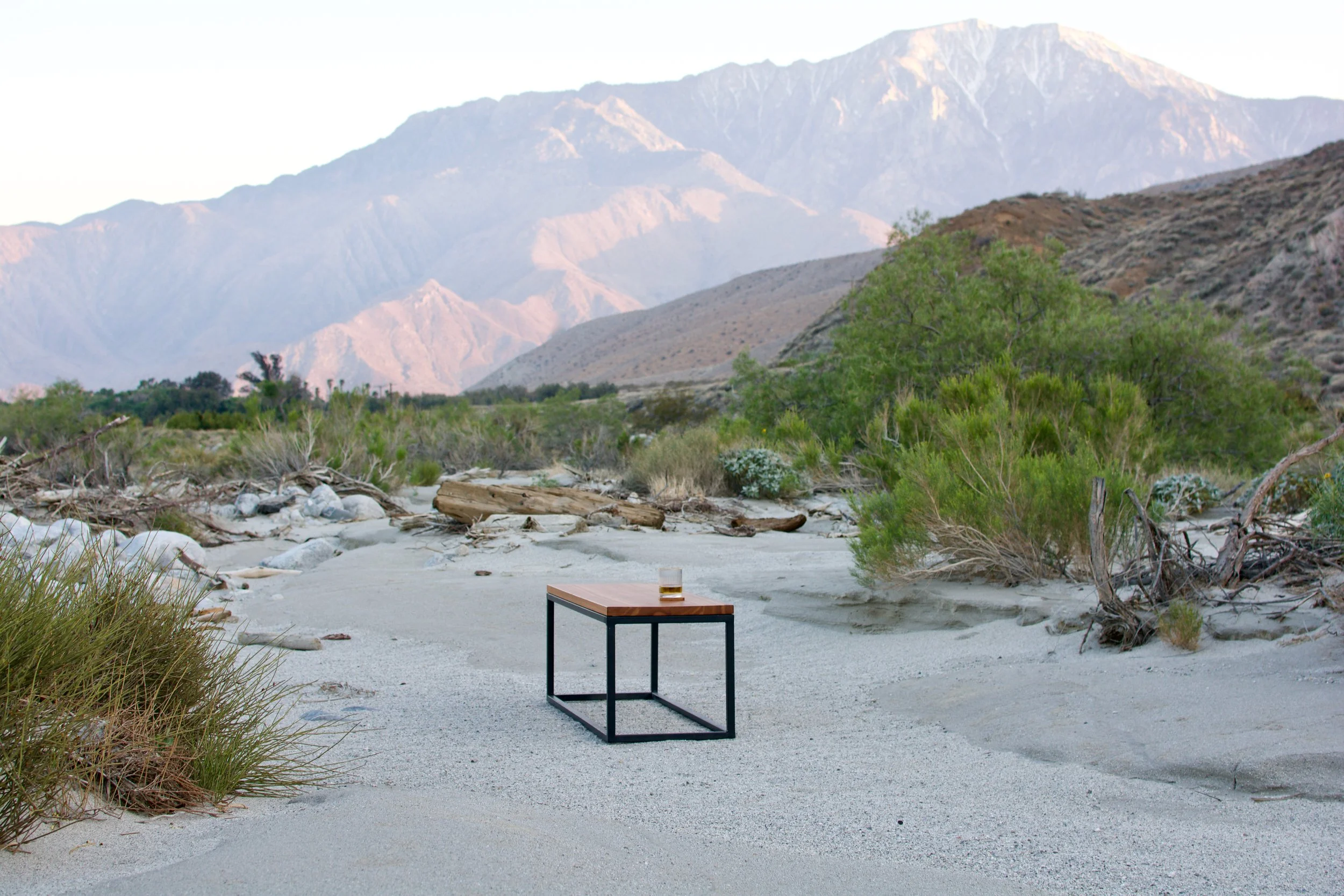A desert landscape with mountains in the background, green shrubs and bushes, a small black metal framed table with a wooden top, and a glass of amber-colored liquid on the table.