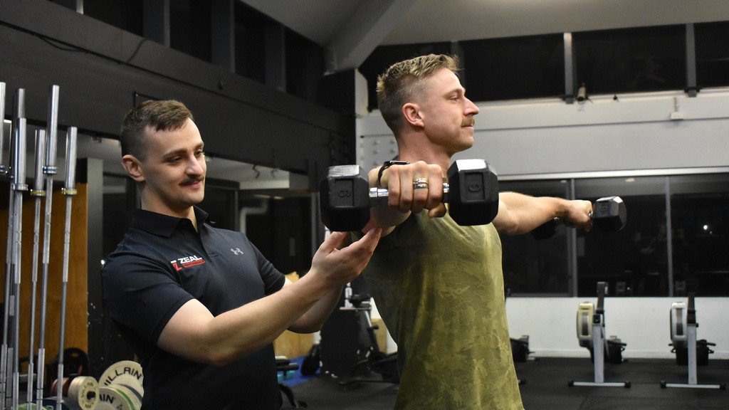 A man in a yellow shirt is lifting a dumbbell while a trainer spotlights him in a gym.