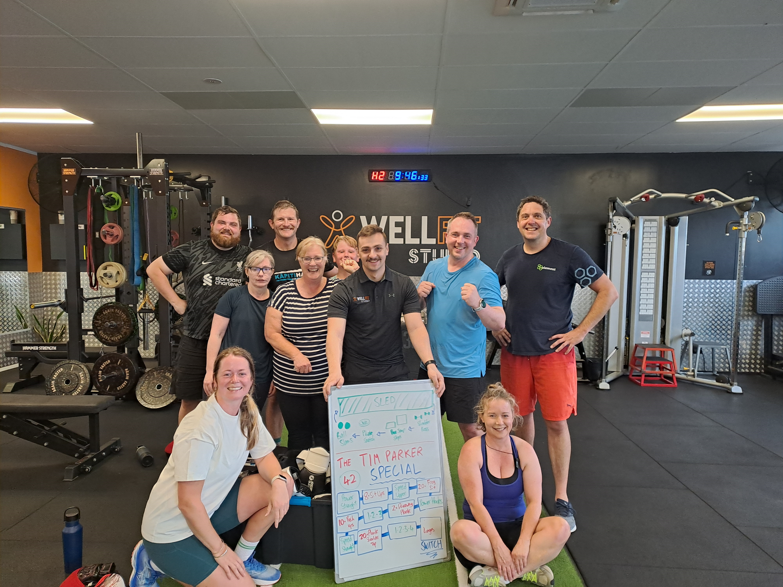 Group of people posing in a gym with fitness equipment and a whiteboard displaying workout info.