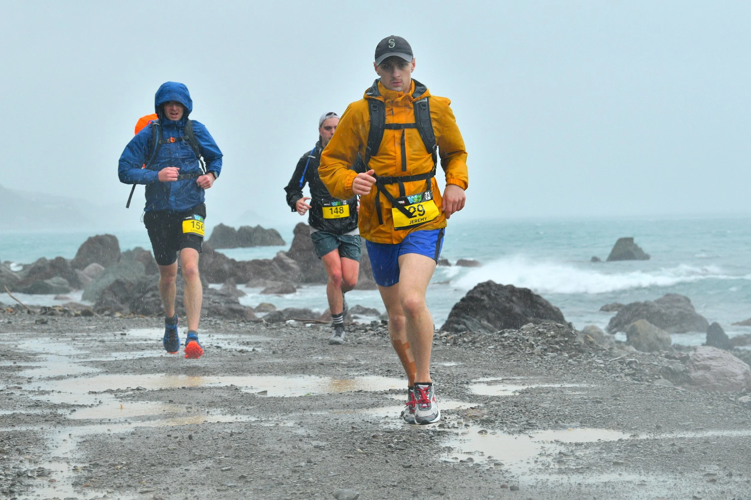 Group of runners participating in a trail race along a rocky and wet shoreline during rainy weather, wearing moisture-resistant clothing and numbered bibs.