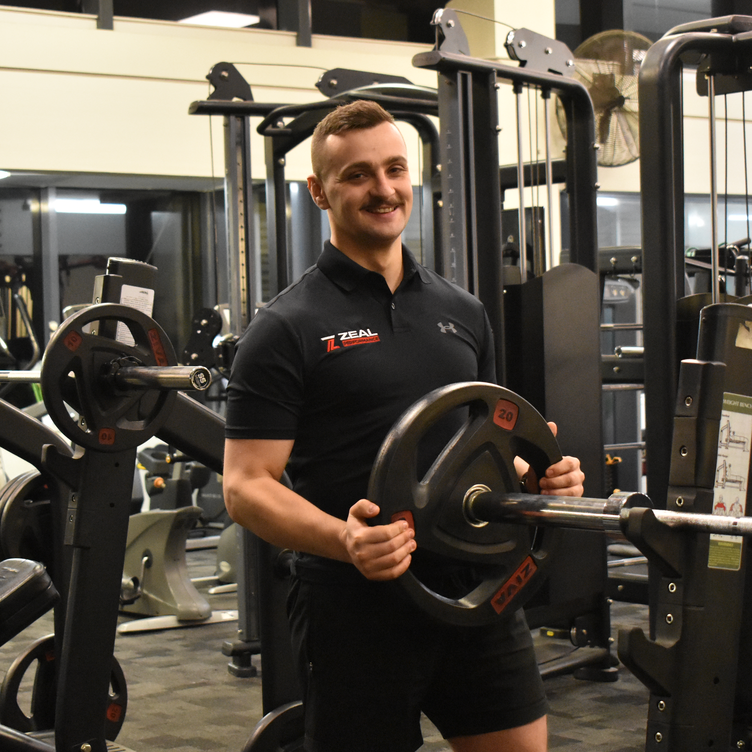 A man holding weight plates in a gym, smiling, wearing a black Under Armour shirt and shorts, surrounded by gym equipment.