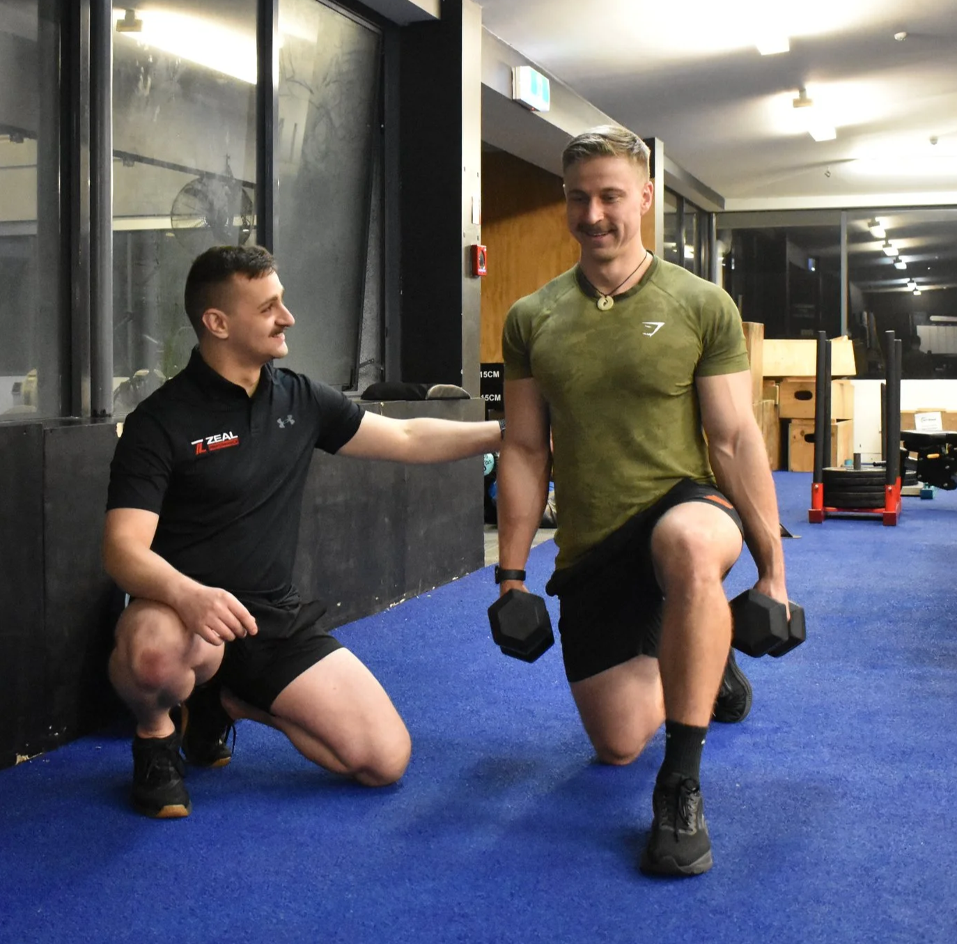 Two men working out in a gym; one is kneeling on the floor with a hand on the other's shoulder, who is holding dumbbells and performing a lunge