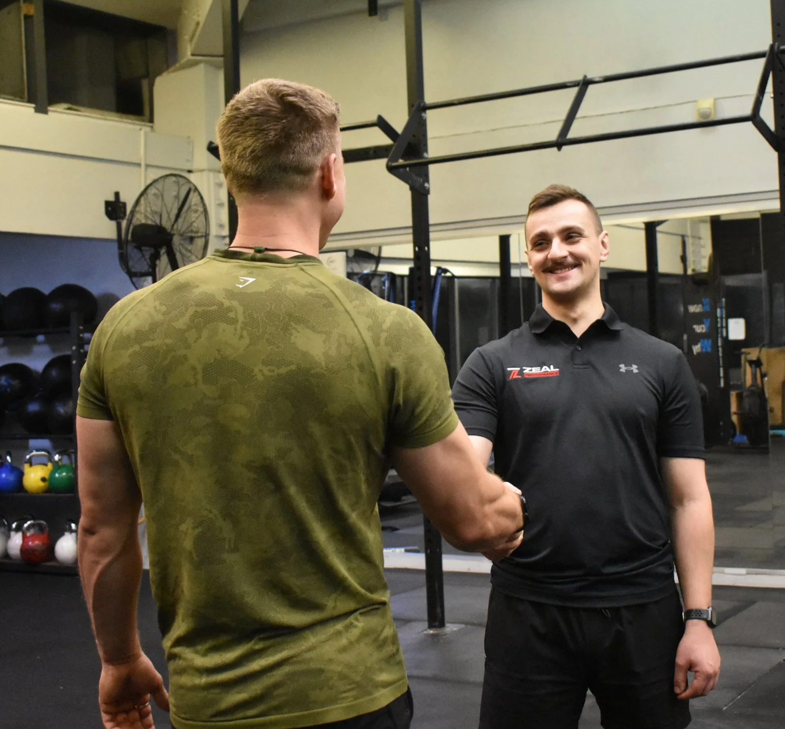 Two men shaking hands in a gym, one wearing a green camouflage shirt and the other in a black polo with logo, smiling, gym equipment in the background.