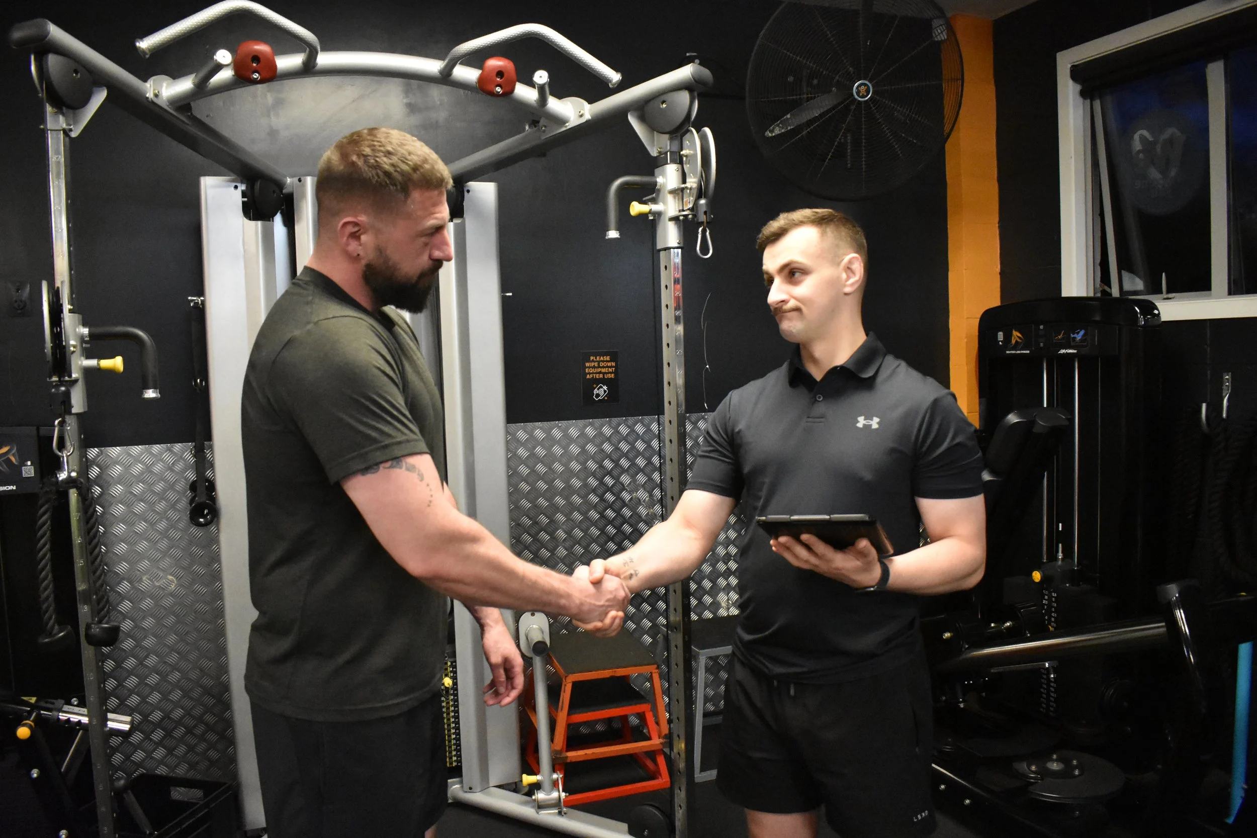Two men shake hands in a gym, one holding a tablet. The gym has black walls, workout equipment, and a large fan on the wall.
