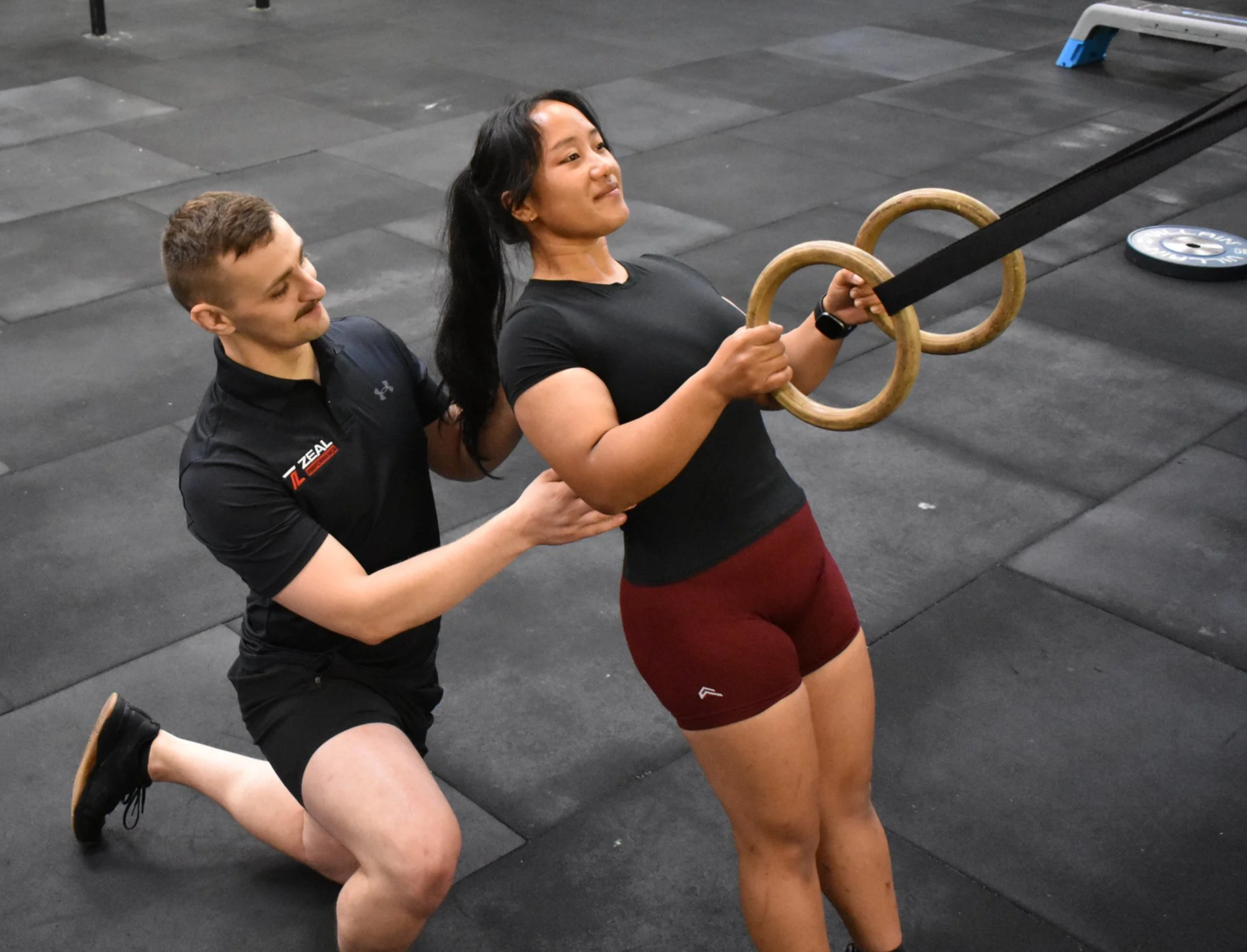 A woman holding gymnastic rings during a workout with a trainer kneeling behind her, guiding her on a black gym floor.