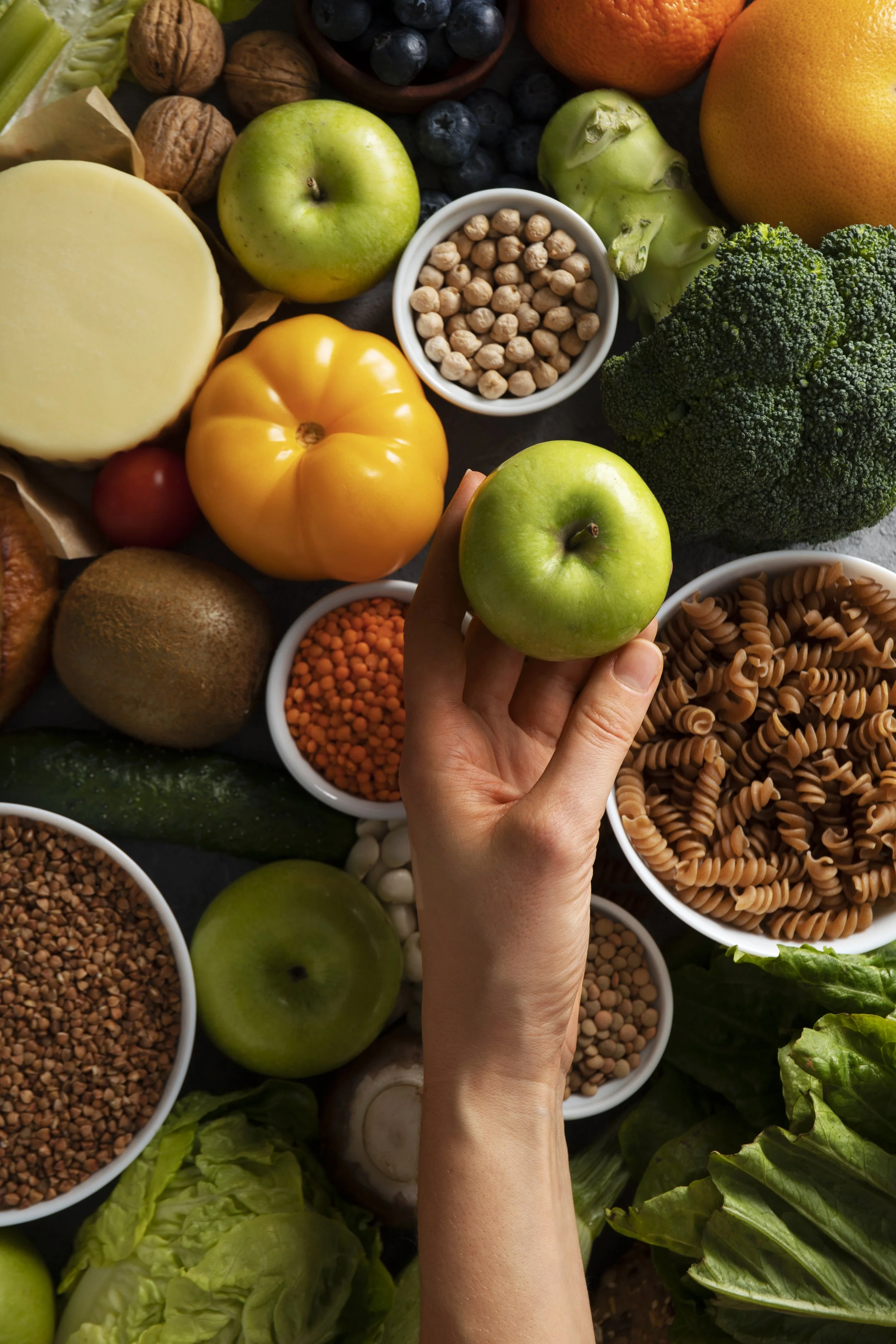 A person holding a green apple over a variety of fresh fruits, vegetables, and grains including broccoli, tomatoes, lettuce, cauliflower, apples, oranges, blueberries, walnuts, chickpeas, lentils, and different types of pasta.