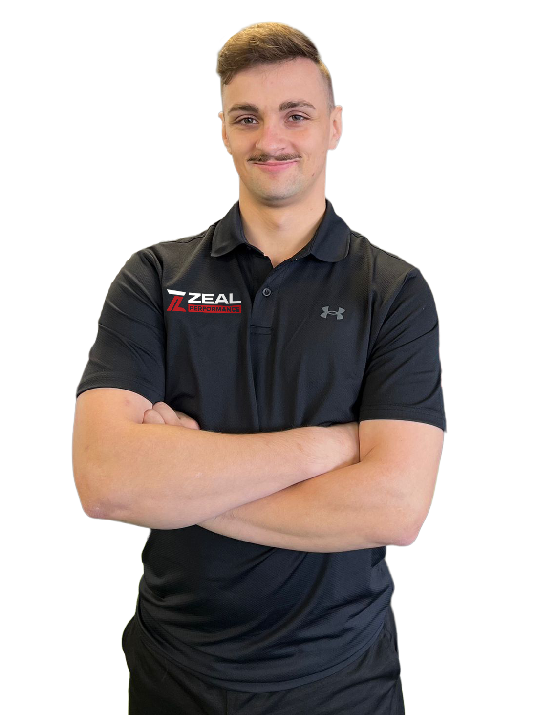 Young man with crossed arms smiling, wearing a black Under Armour shirt with Zeal Performance logo, in a gym with black walls and gym equipment in the background.