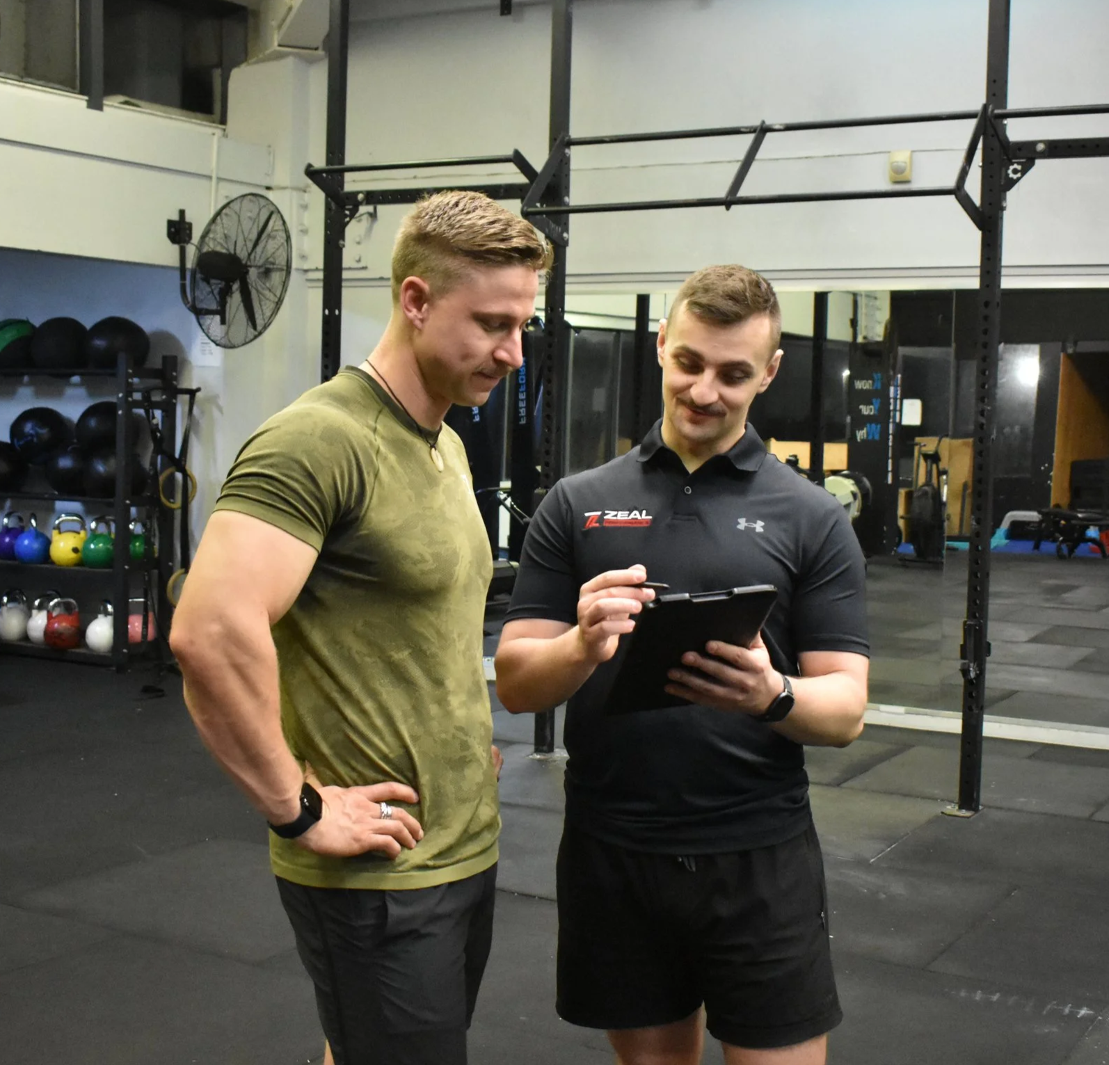 Two men in athletic clothing standing in a gym, looking at a clipboard or tablet together, while discussing something. The gym has weights, kettlebells, and fitness equipment in the background.