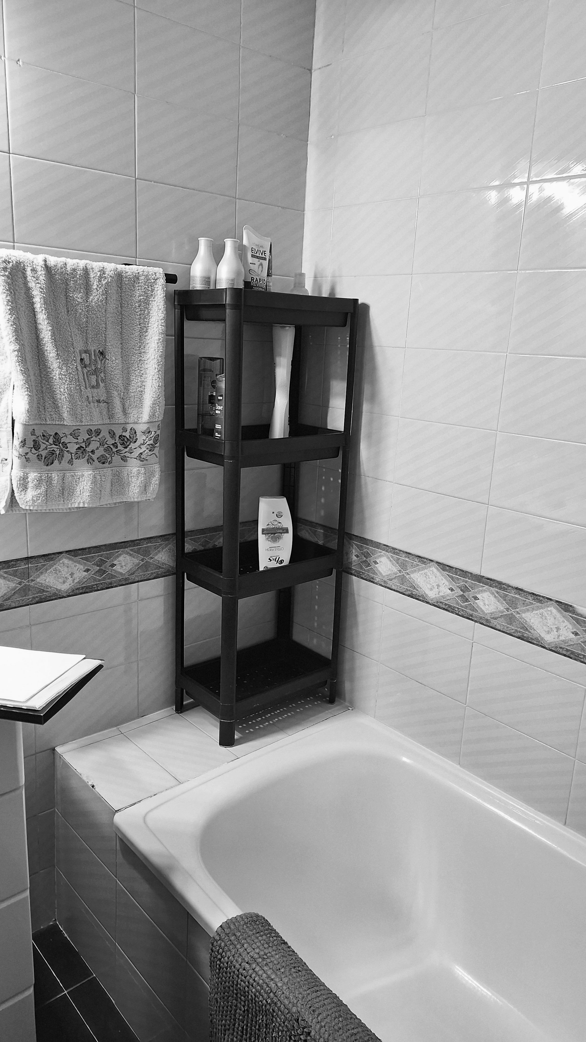 A bathroom featuring a bathtub, a black shelving unit, and a towel hanging on a rail. The shelf holds various bottles and a box of Elvive hair products.