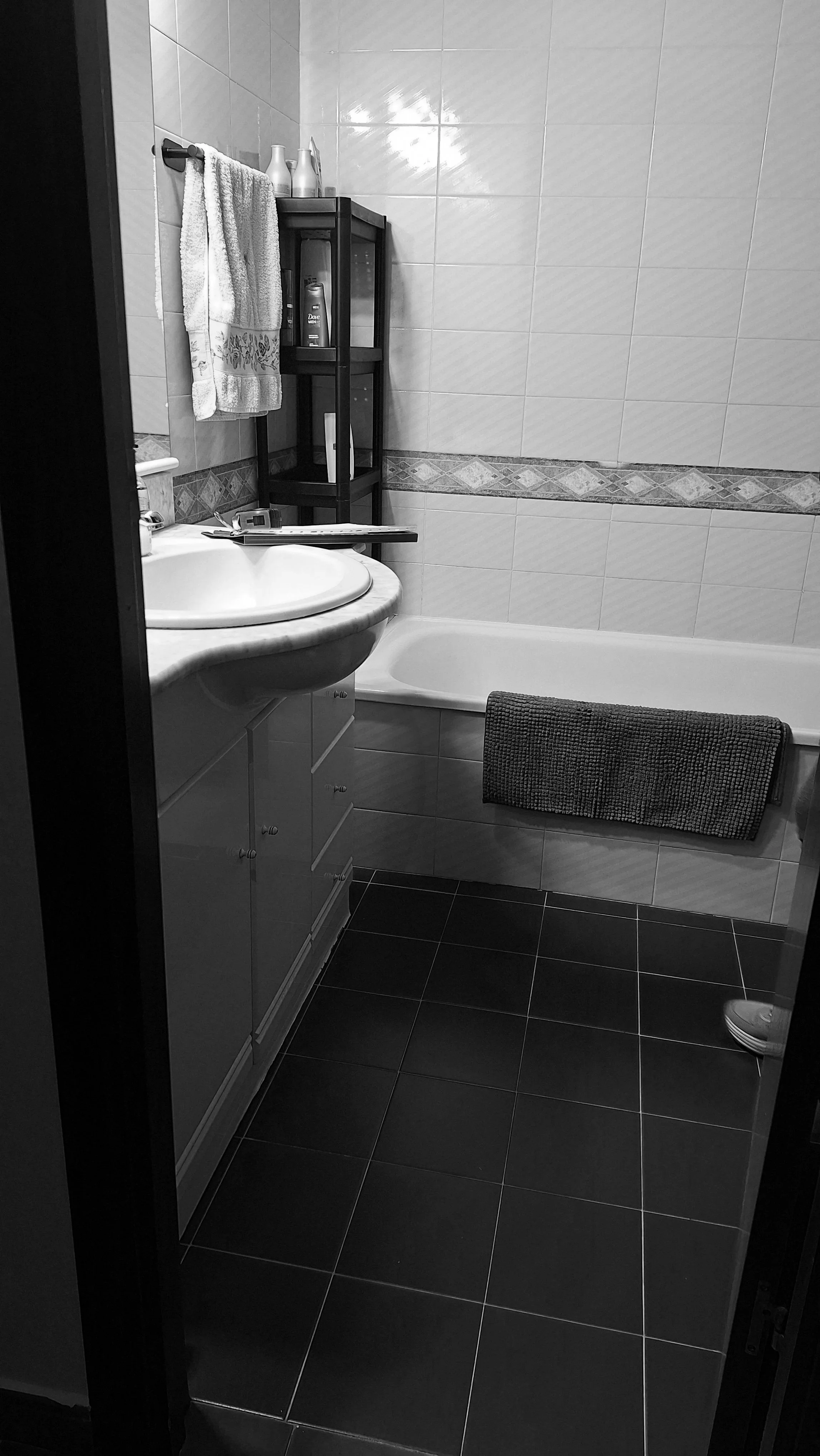 A small bathroom with a tiled wall and floor, a sink with storage drawers below, a towel hanging on a rack, a black shelf with grooming products, a bathtub with a textured bath mat, and a pair of shoes partially visible