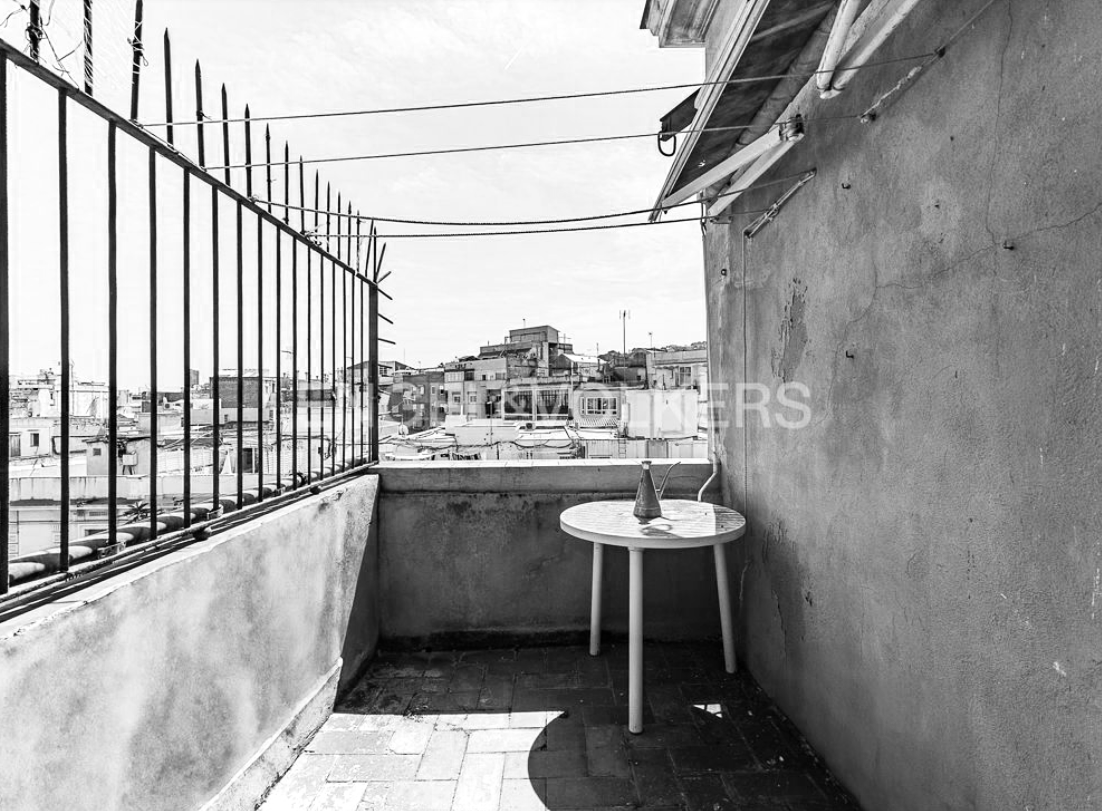 Black and white photo of a small rooftop balcony with a table and a bottle on it, overlooking a cityscape with multiple buildings and rooftops.