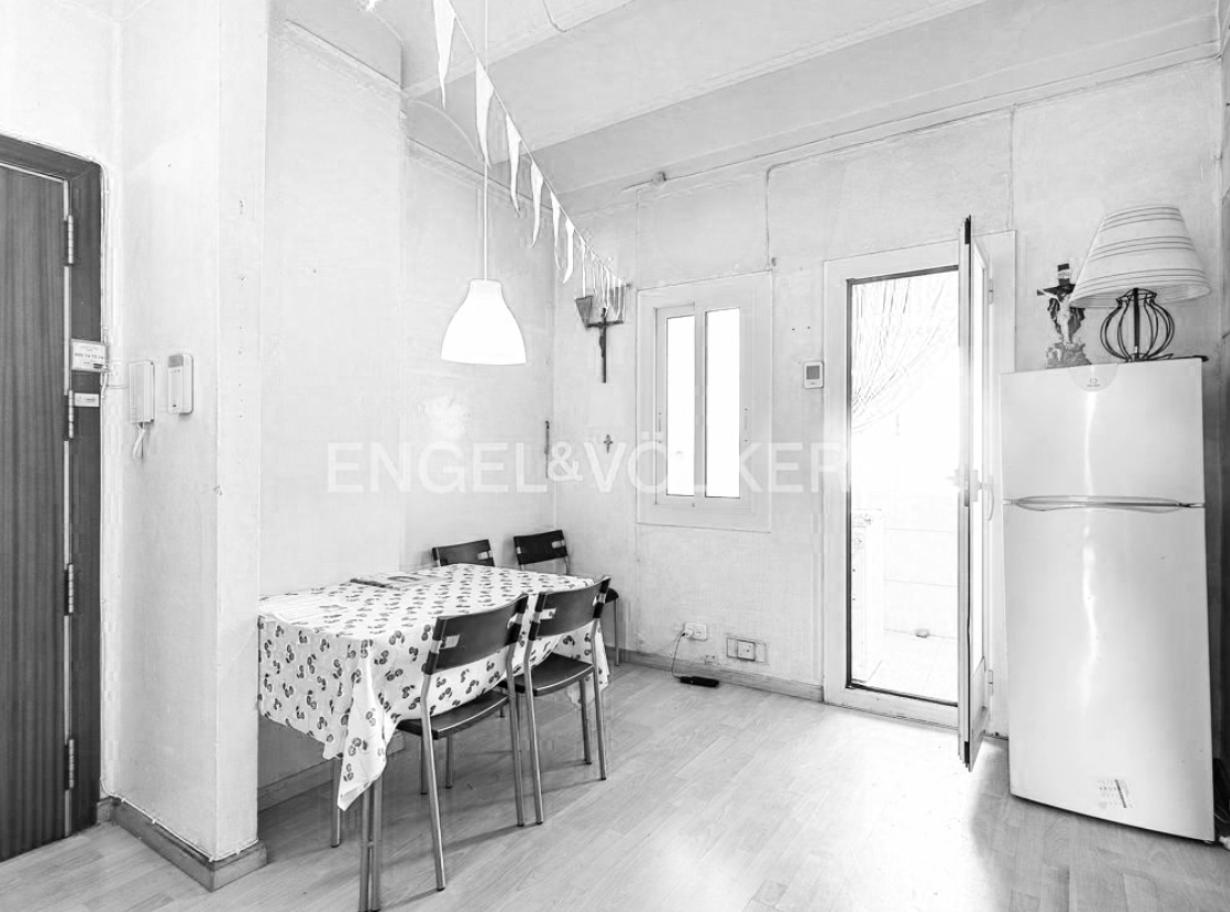 Interior of a small kitchen or dining area with a table covered by a patterned tablecloth, four chairs, a white refrigerator, a window, a door, and a pendant light hanging from the ceiling.