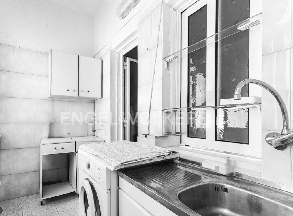 Black and white photo of a small kitchen with a washing machine below a countertop, a sink, and a window with clotheslines. Cabinets and a small table are also visible.