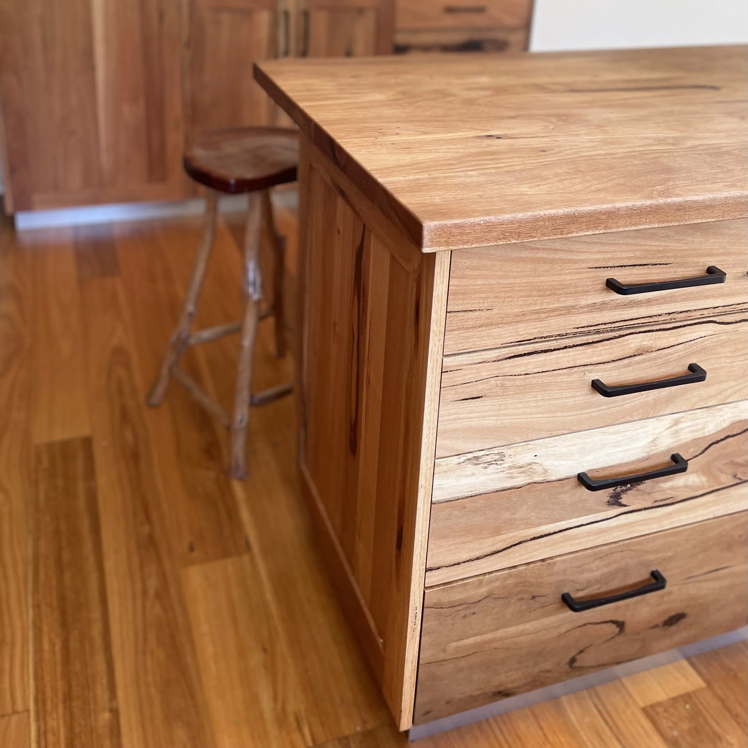 A wooden kitchen island with four drawers and black handles, next to a rustic wooden stool with natural legs, on a wooden floor.