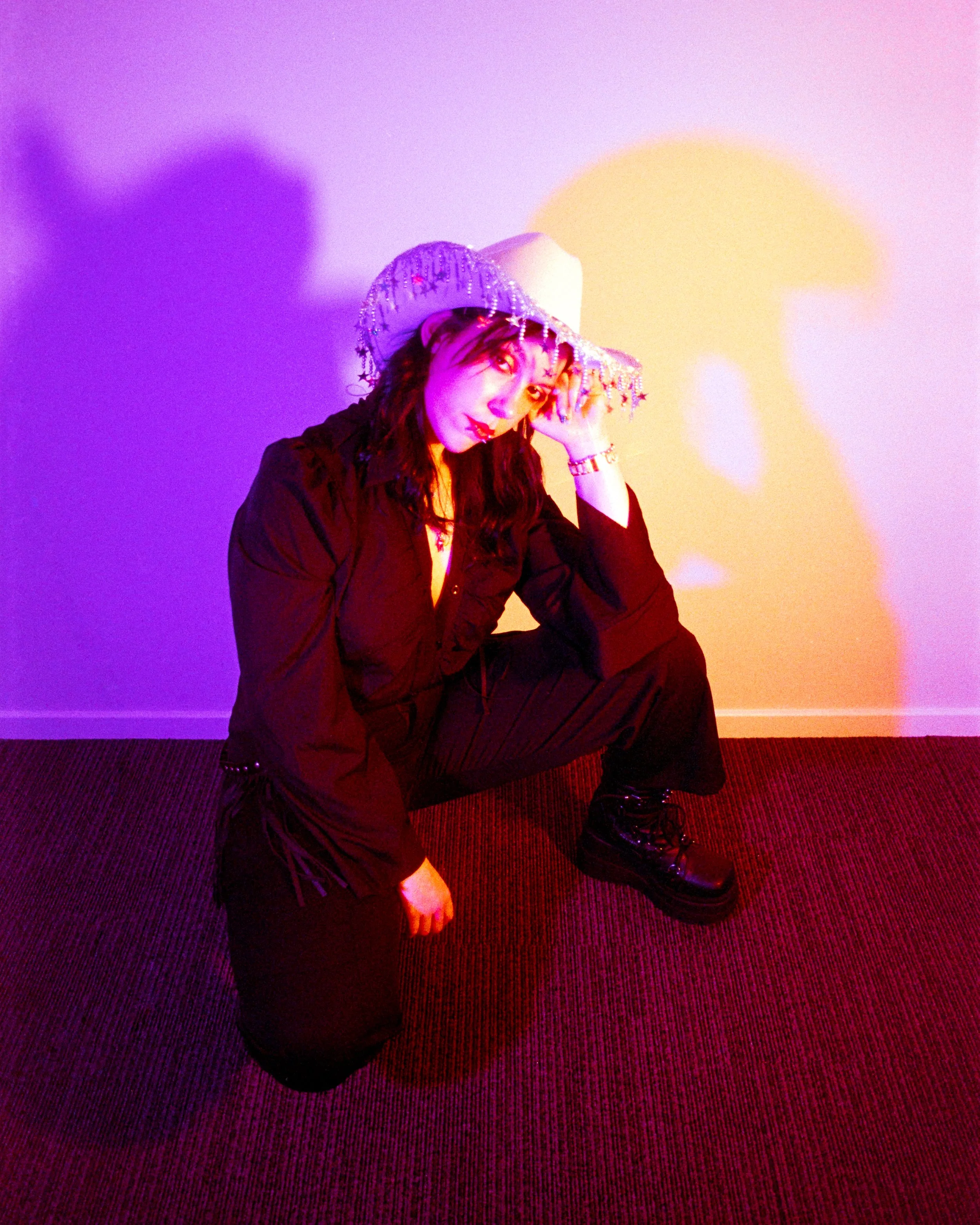 Self portrait of Josie in her Rhinestone Cowgirl outfit, pink and peach lights illuminating her, kneeling on the ground and resting her head on one hand, on 35mm Film - Ultramax