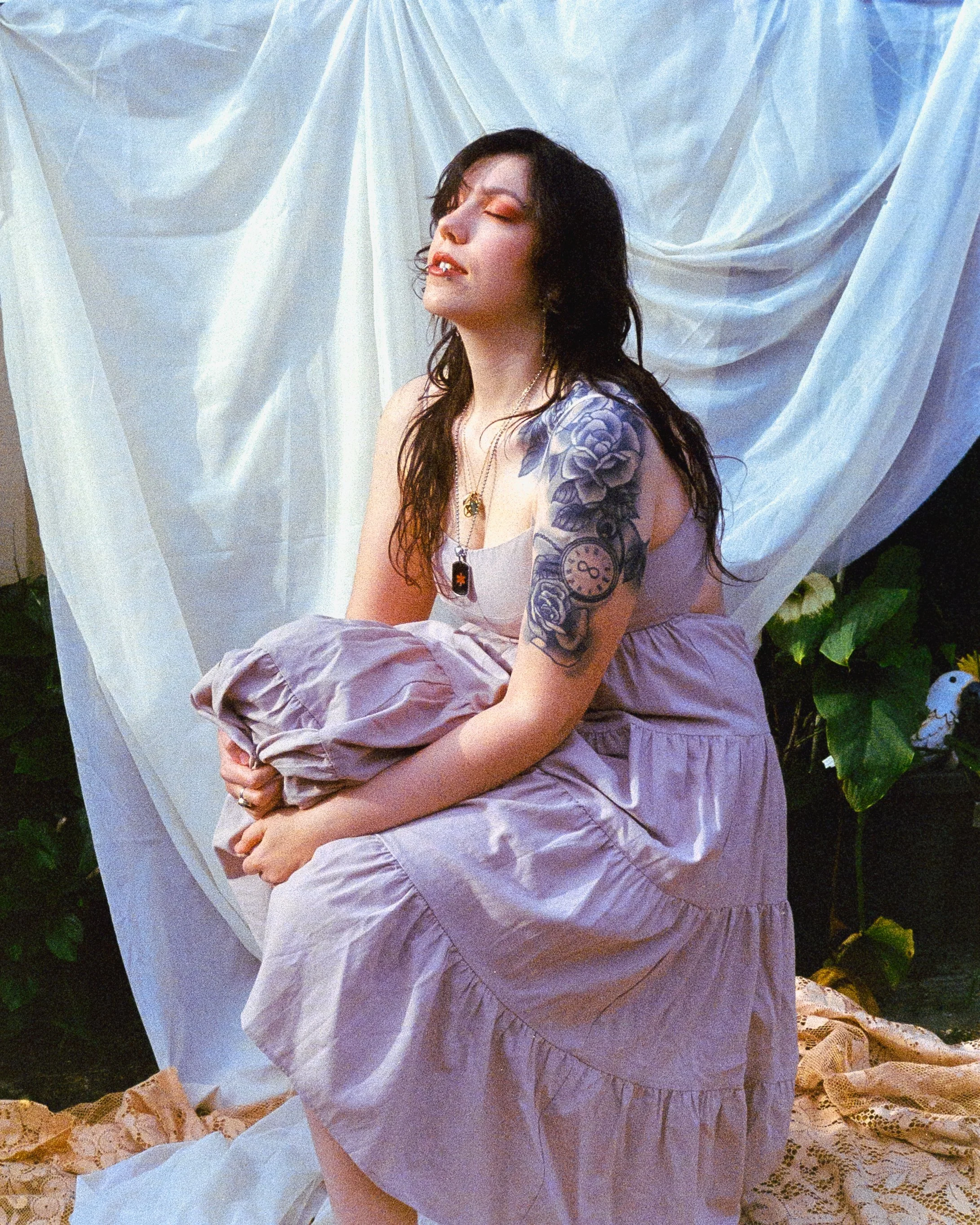 A self portrait of Josie in the backyard with a simple sheet draped over a plant for a backdrop and lace around her feet. She's wearing a beige dress and basking in the sun, on 35mm Film - Ultramax