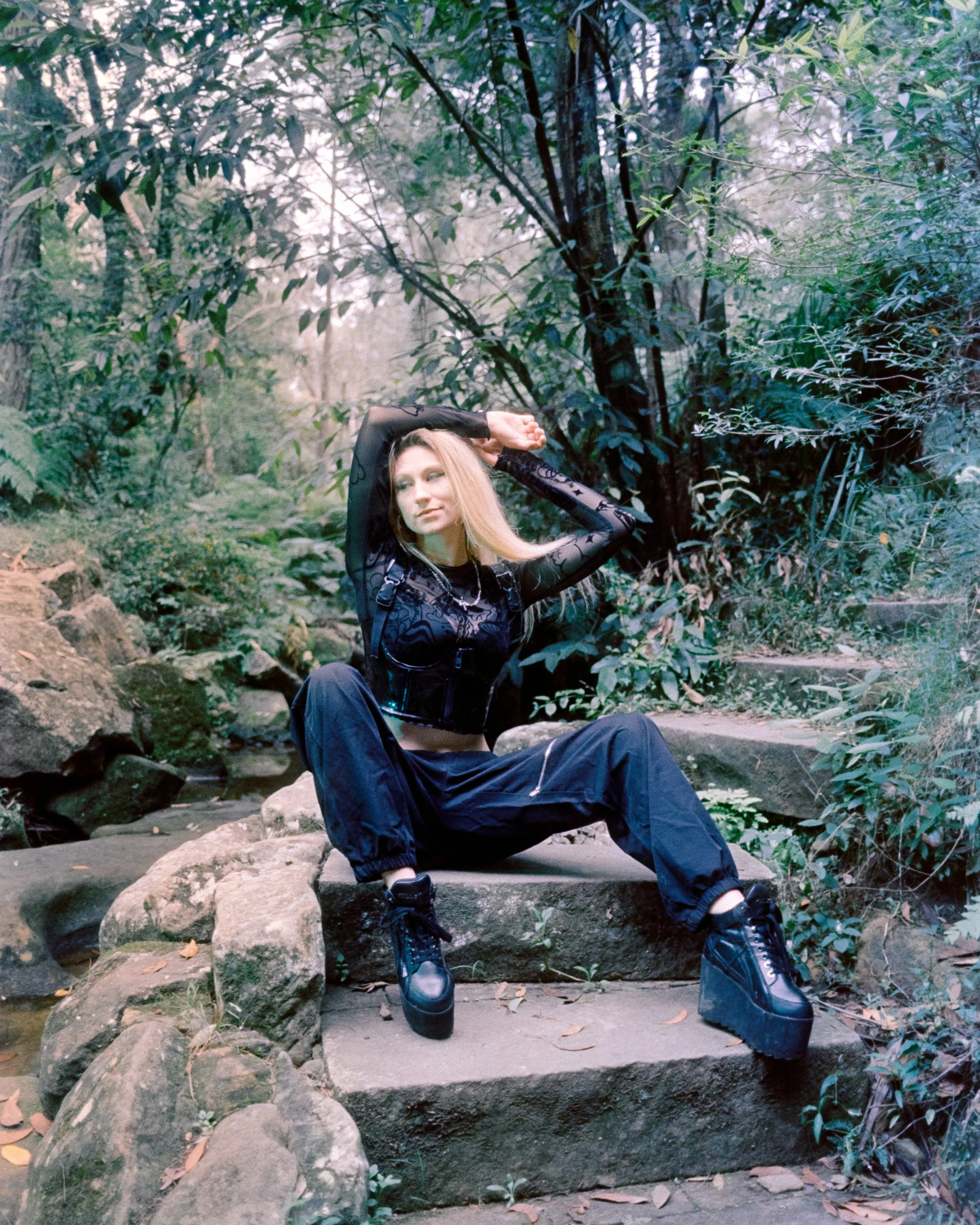 An image of a gothic alternative woman in the forest, sitting on some stone steps with her arms above her head, looking over her shoulder, lit up by green lighting, shot on 35mm Film - 200T Cinefilm