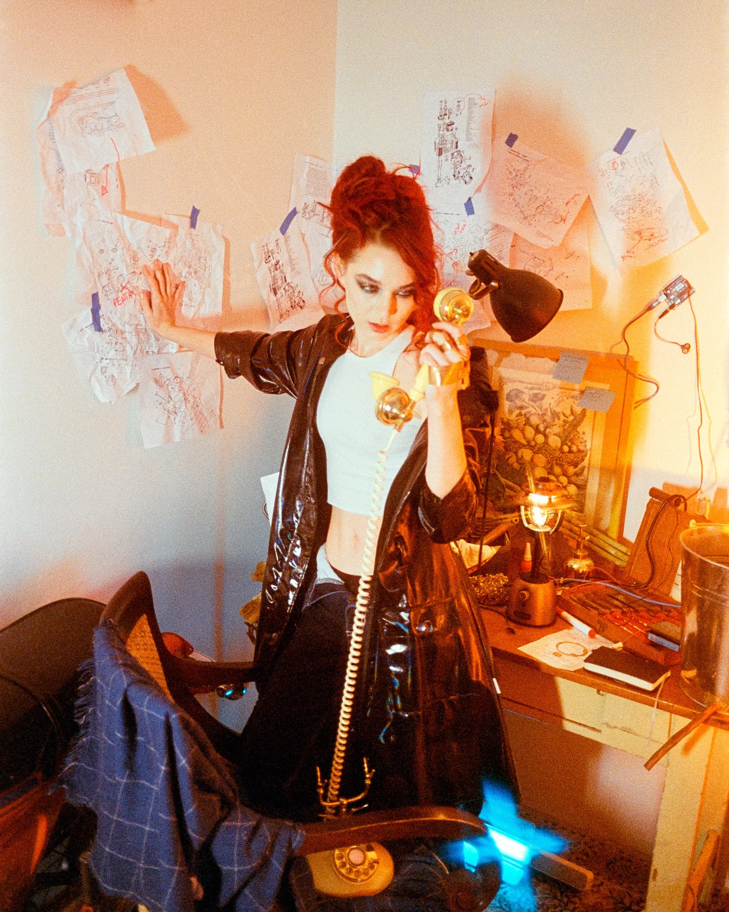 A woman at her workbench, looking frustrated at an old phone, hand on the wall full of schematics, illuminated by orange lights, on 35mm Film - Ultramax
