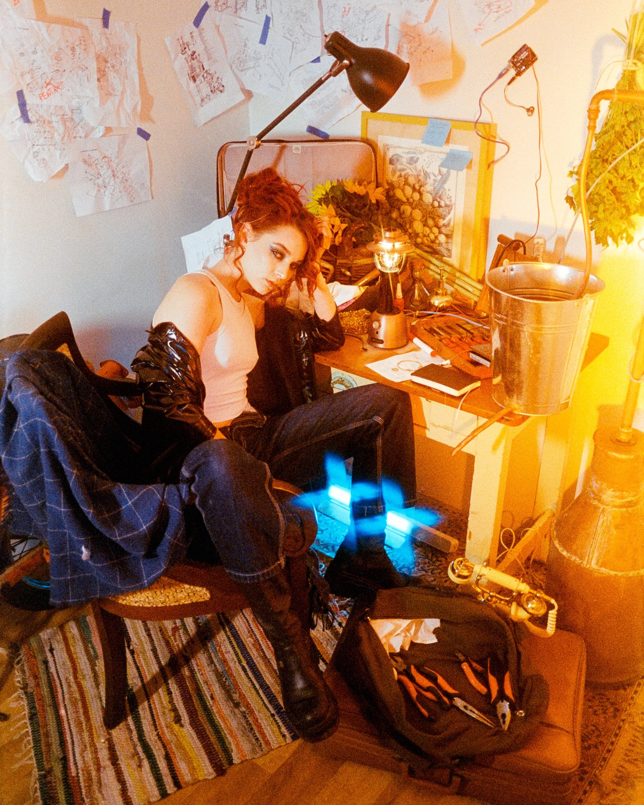 a woman sitting at a workbench covered in projects and supplies, multuiple glowing lamps on the desk, and she looks at the camera, on 35mm Film - Ultramax
