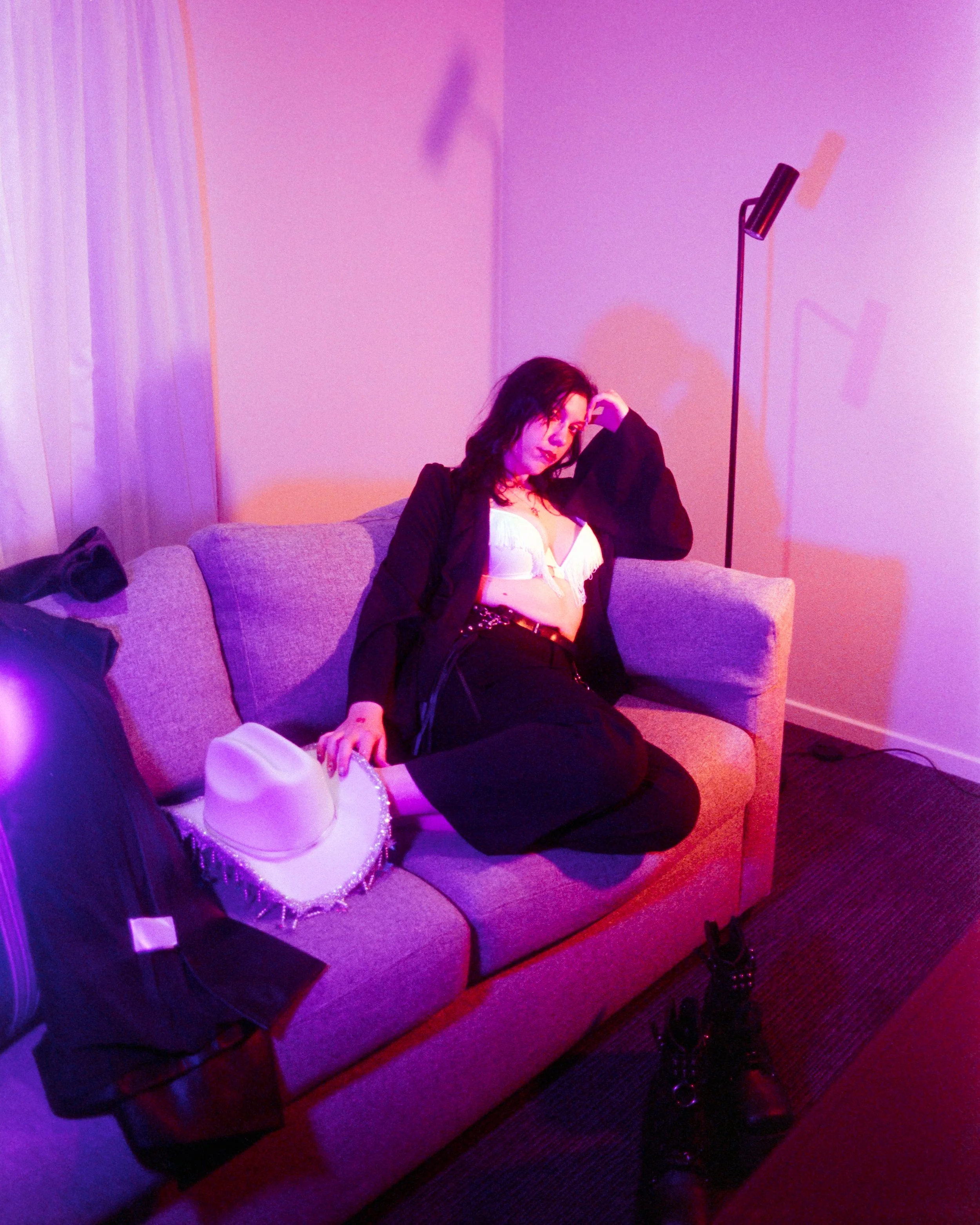 Self portrait of Josie in her Rhinestone Cowgirl outfit, pink and peach lights illuminating her, she's on the couch with her legs tucked up, head resting on her hand with a soft smile, on 35mm Film - 400D Cinefilm