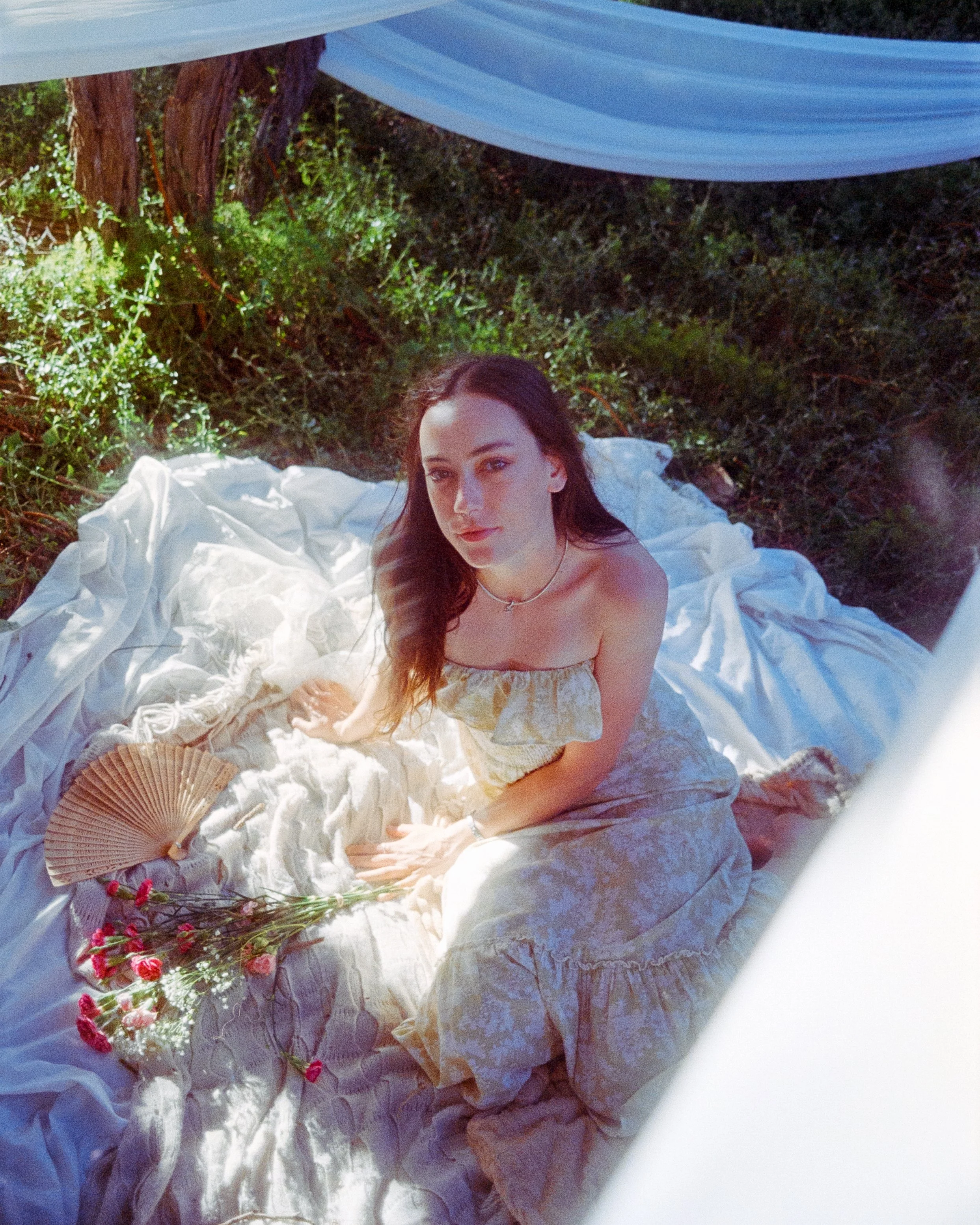 a dynamic image of a woman looking up at the camera while sitting on draped fabric with flowers and a handheld fan on the ground near her, golden sunlight shining, on 35mm Film - Ultramax
