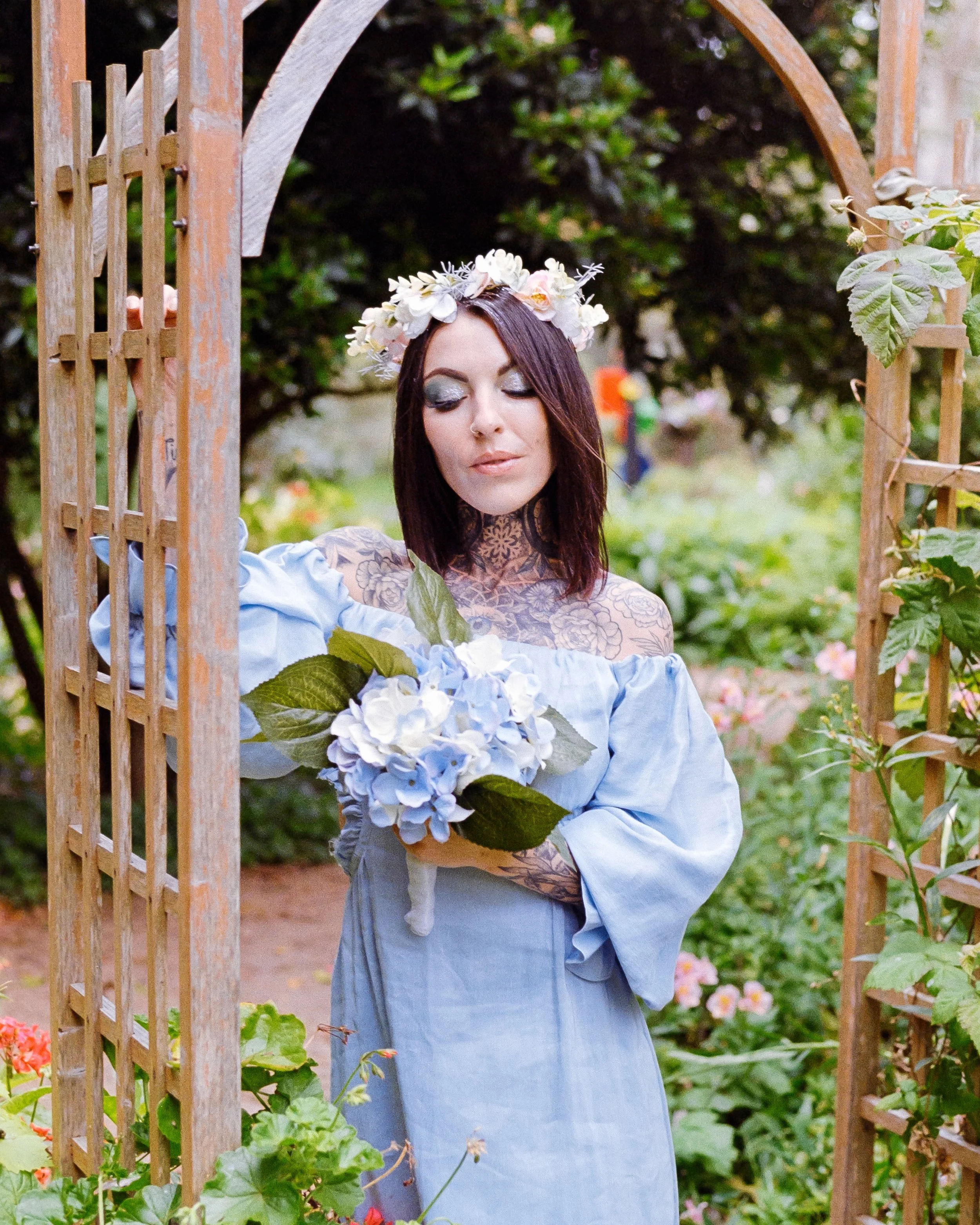 a tattooed woman wearing a baby blue dress, holding flowers and with a flower crown in her hair, leaning on a trellis in a garden, on 35mm Film - Pro Image 100