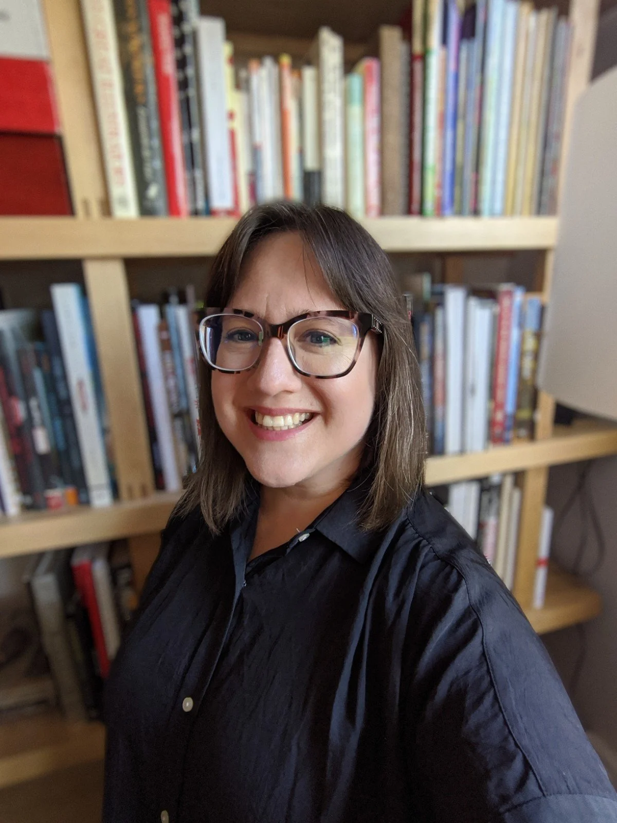 A woman with shoulder-length brunette hair, glasses, and a big smile, standing in front of wooden bookshelves filled with books.